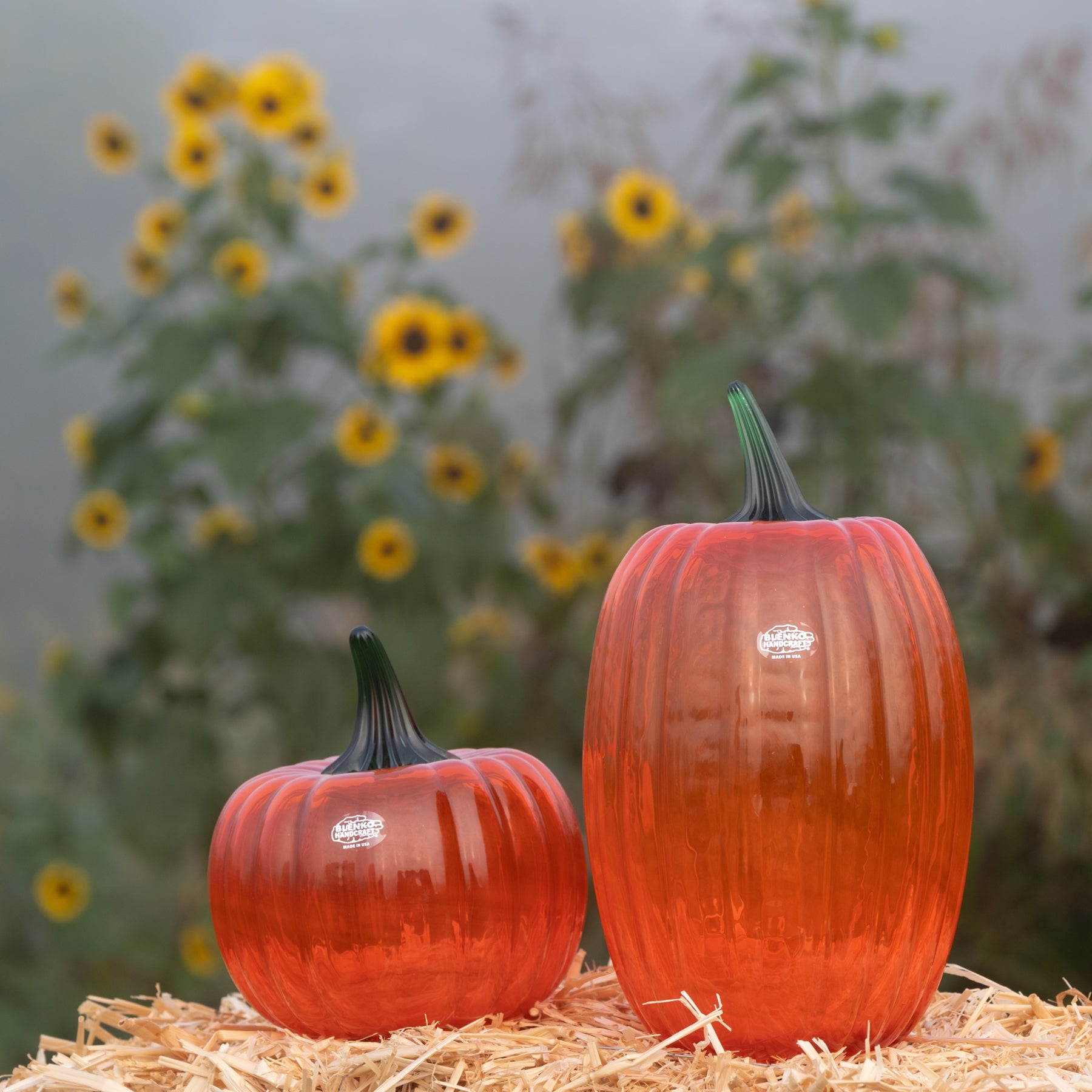 Two orange-red glass pumpkins with dark green stems sit on a bed of straw in front of a blurred background of sunflowers and foliage.