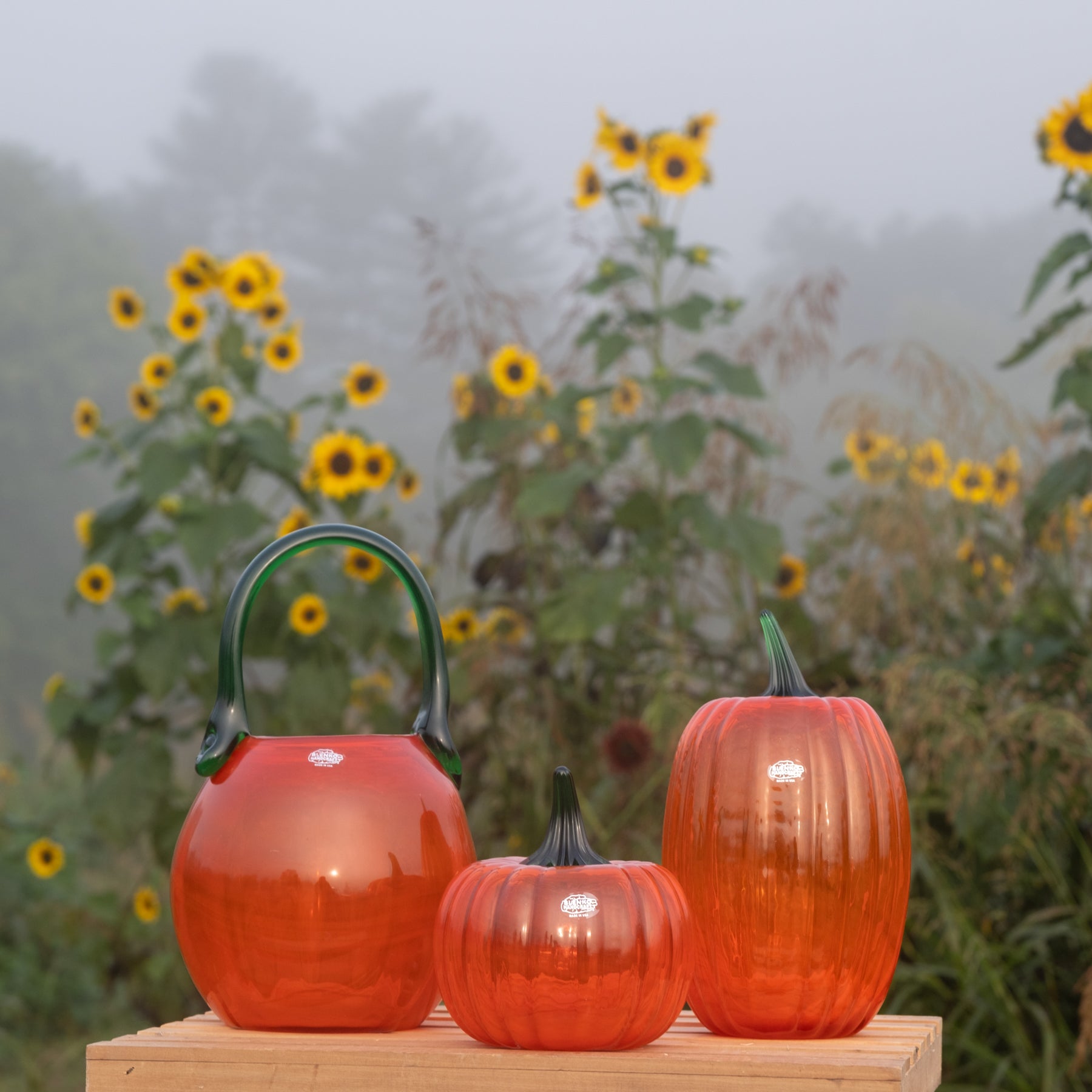 Three translucent orange glass pumpkins in different sizes displayed on a wooden block, with a misty sunflower field in the background.