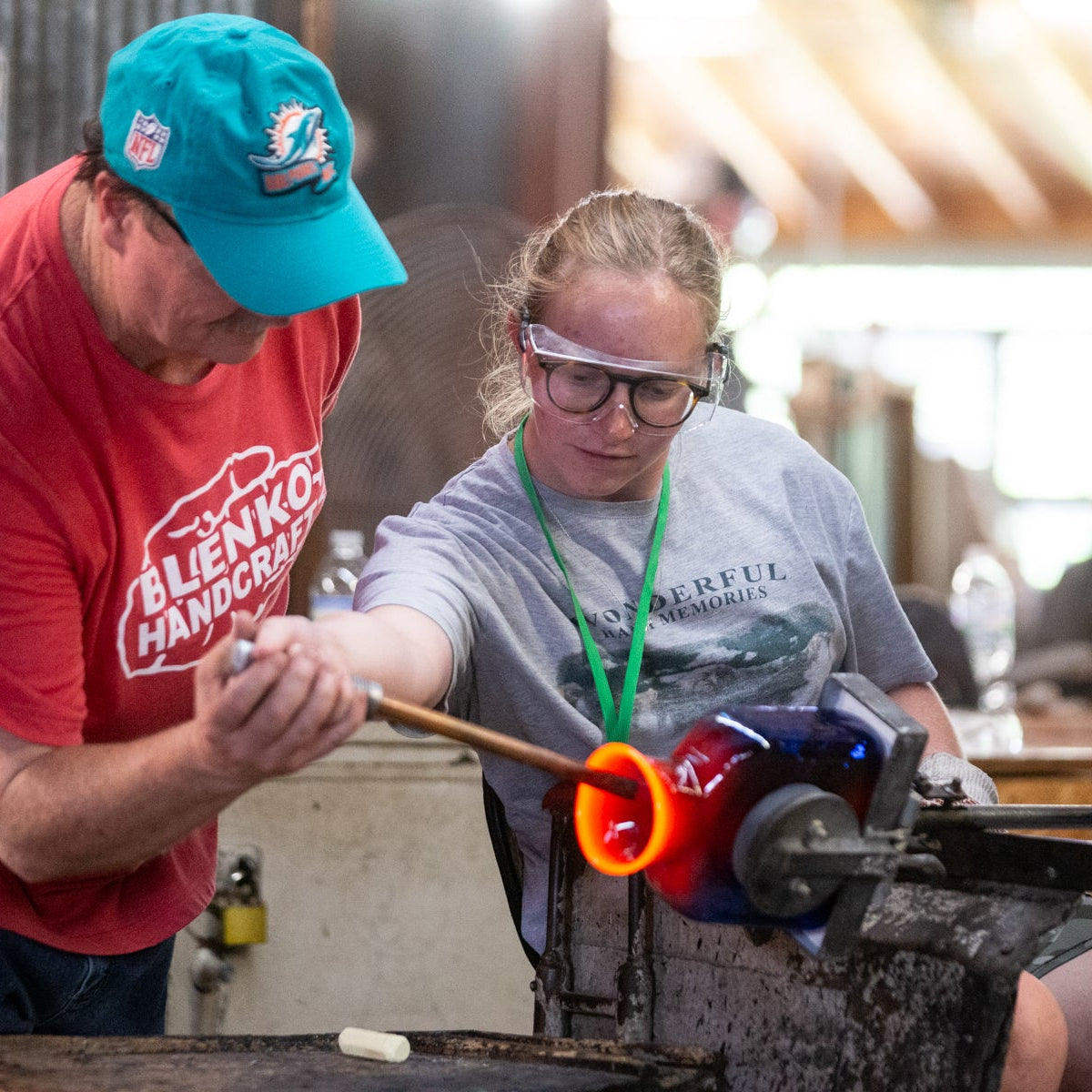 Two people working with glassblowing tools in a workshop setting.