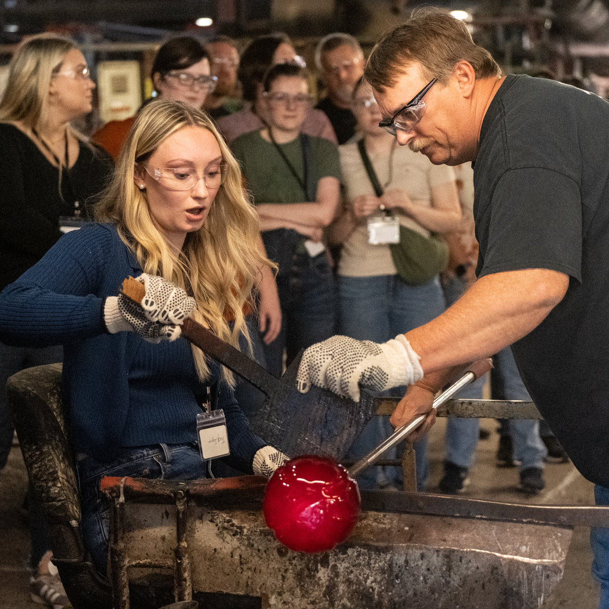 A glassblower shapes a bright red glass bubble over a furnace while a woman watches closely. Several other people stand in the background inside what appears to be a glass studio or workshop.