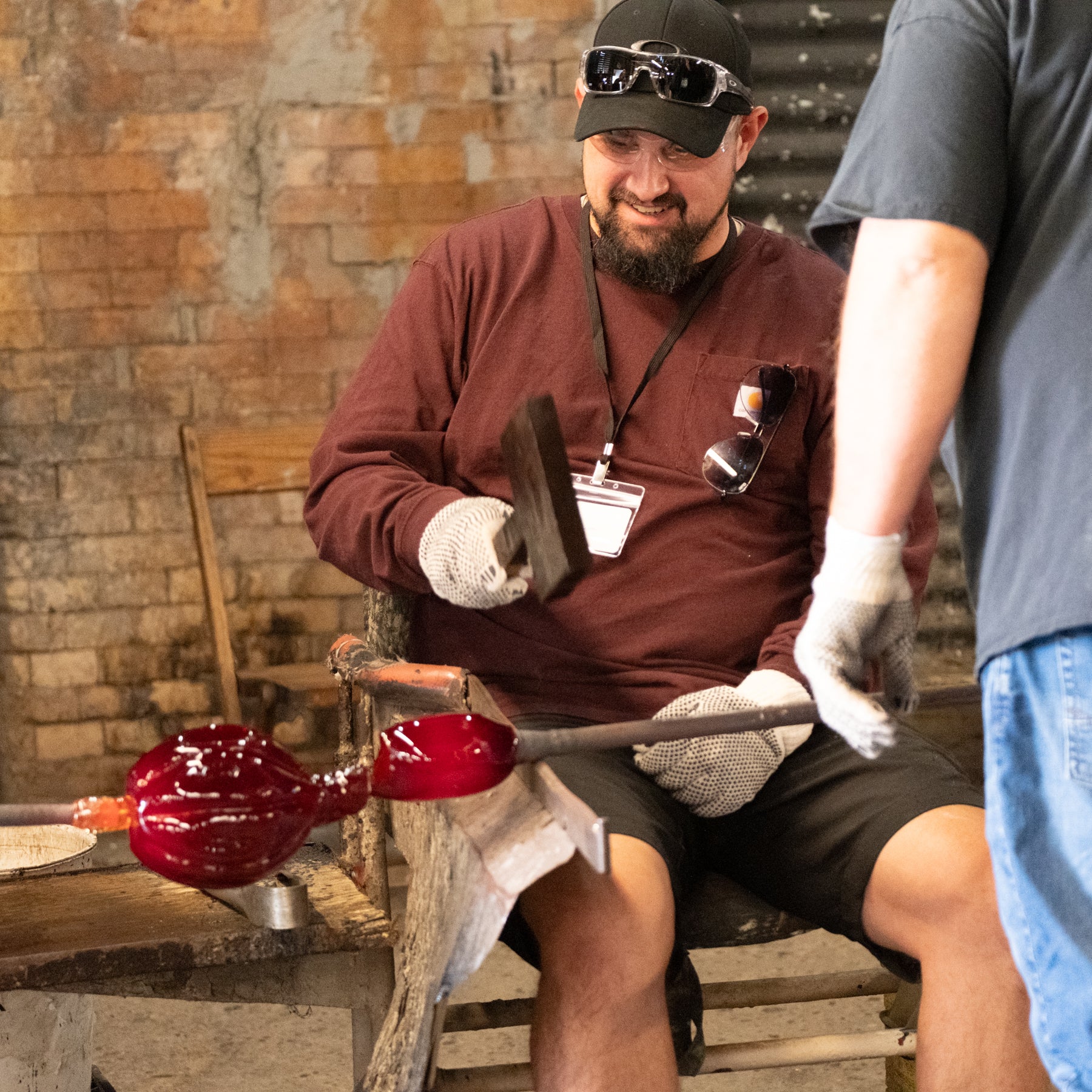 A glassblower in a workshop wearing a maroon long-sleeve shirt, black cap, and white gloves, shaping a red glass bubble on a blowpipe. Another person in a gray shirt is partially visible on the right.