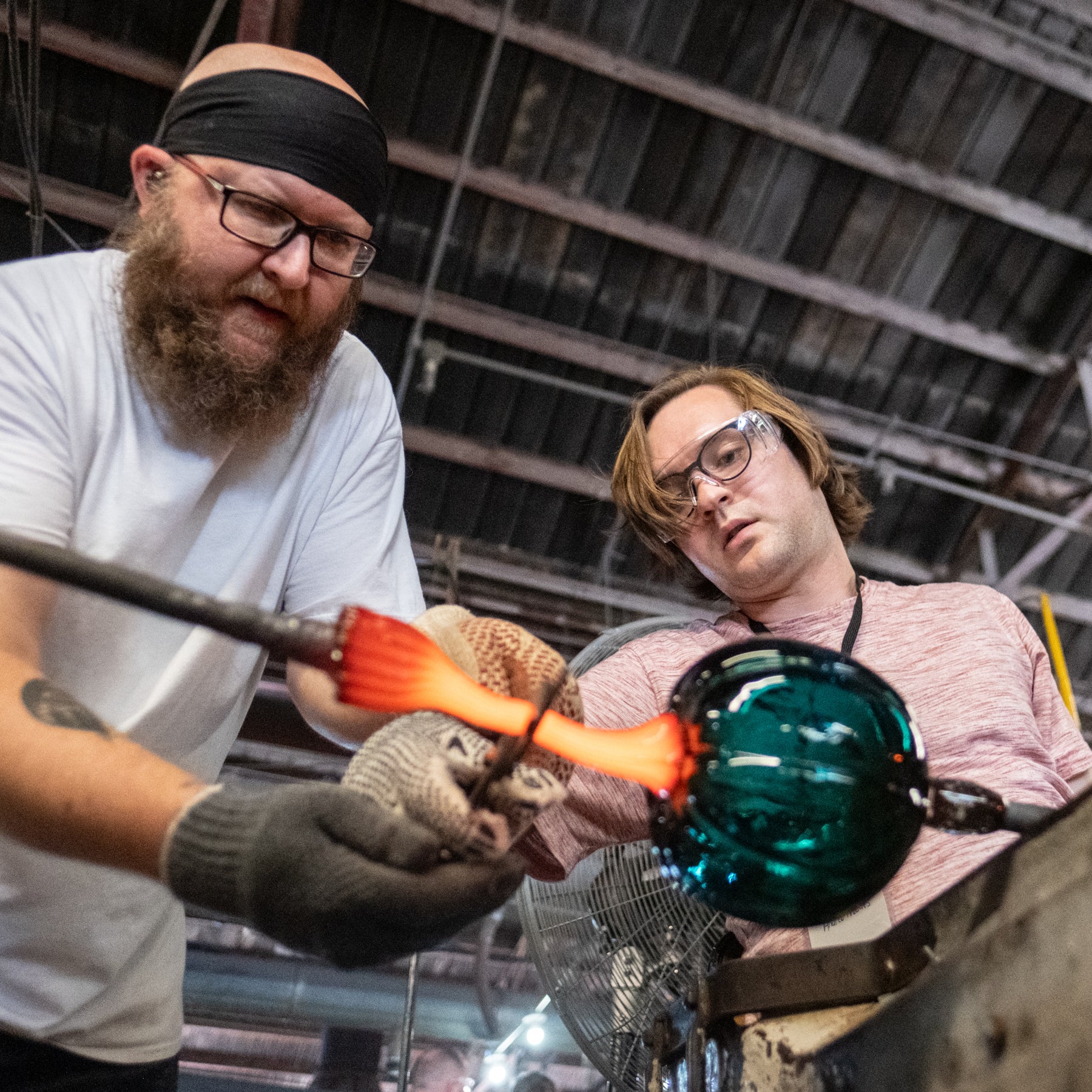 Two glassblowers working in a glassblowing studio. One man in the foreground holds a heated glass rod with a glowing orange-red tip and a dark green glass bubble, while another man watches closely in the background.