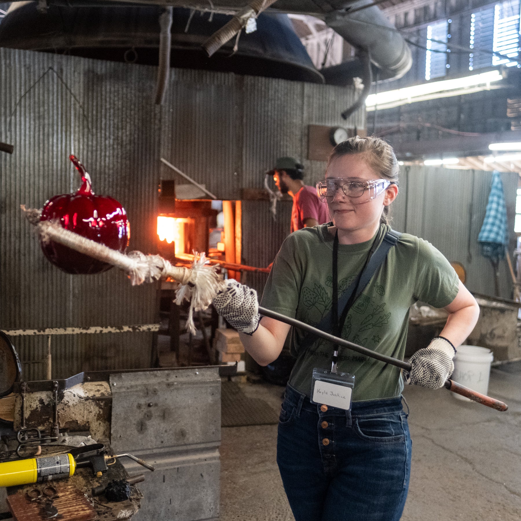 A person in a glassblowing workshop holds a long metal rod with a red glass object attached, standing in front of a furnace. The workshop has corrugated metal walls, tools, and another person in the background.