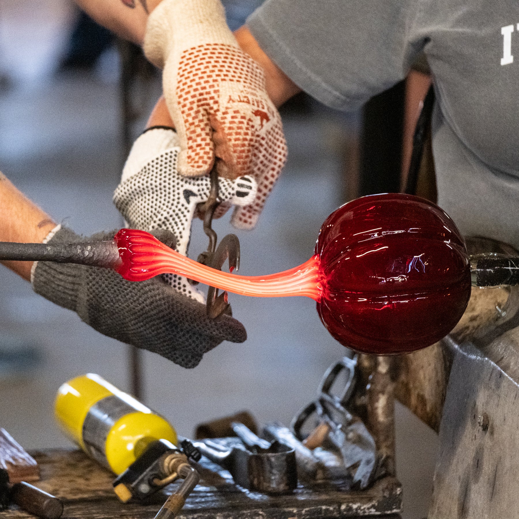 A close-up of a glassblower shaping a glowing red glass bubble with tools and a torch in a workshop setting.