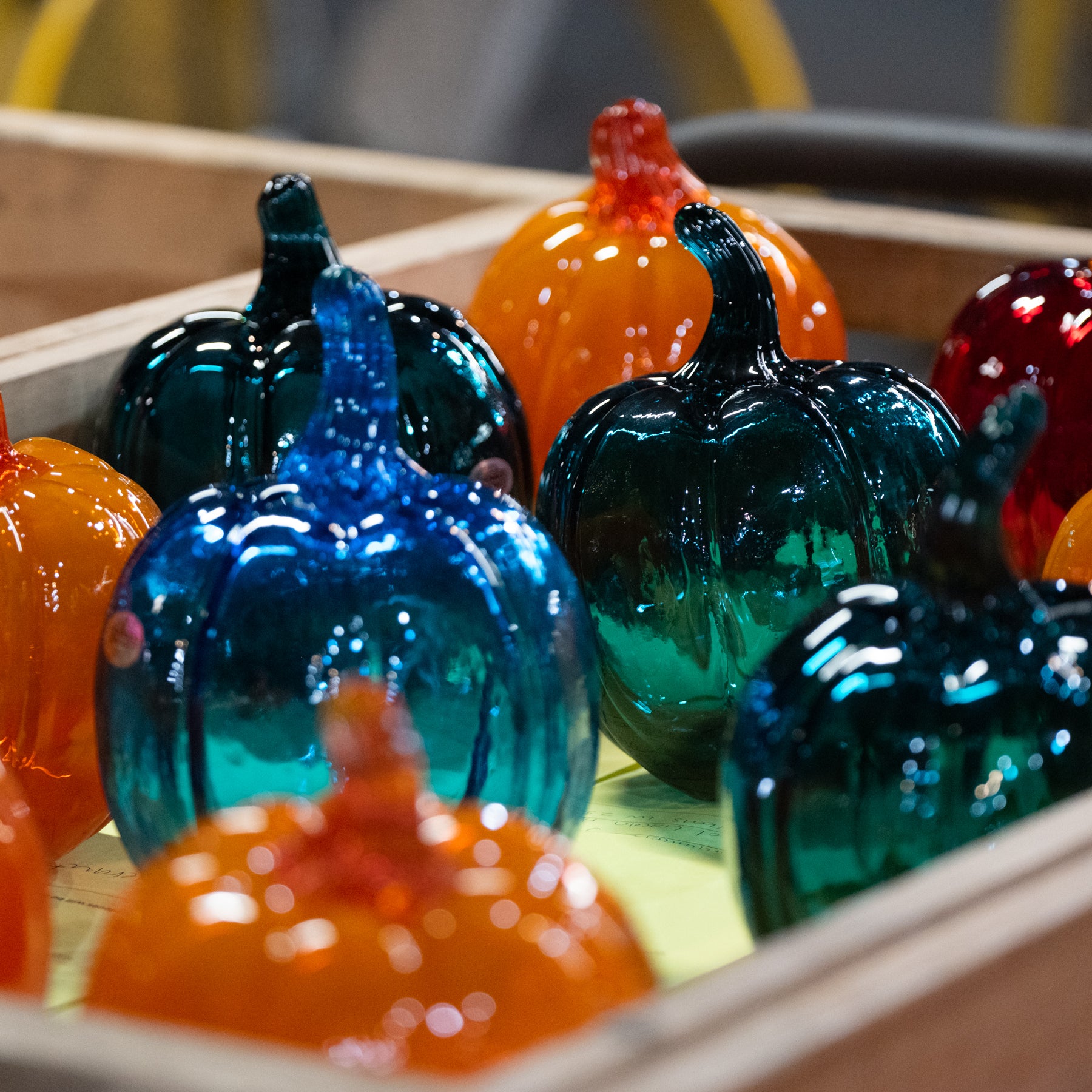 A close-up of glossy, translucent glass pumpkins in autumn colors, including blue, teal, orange, red, and dark green, arranged in a wooden tray.