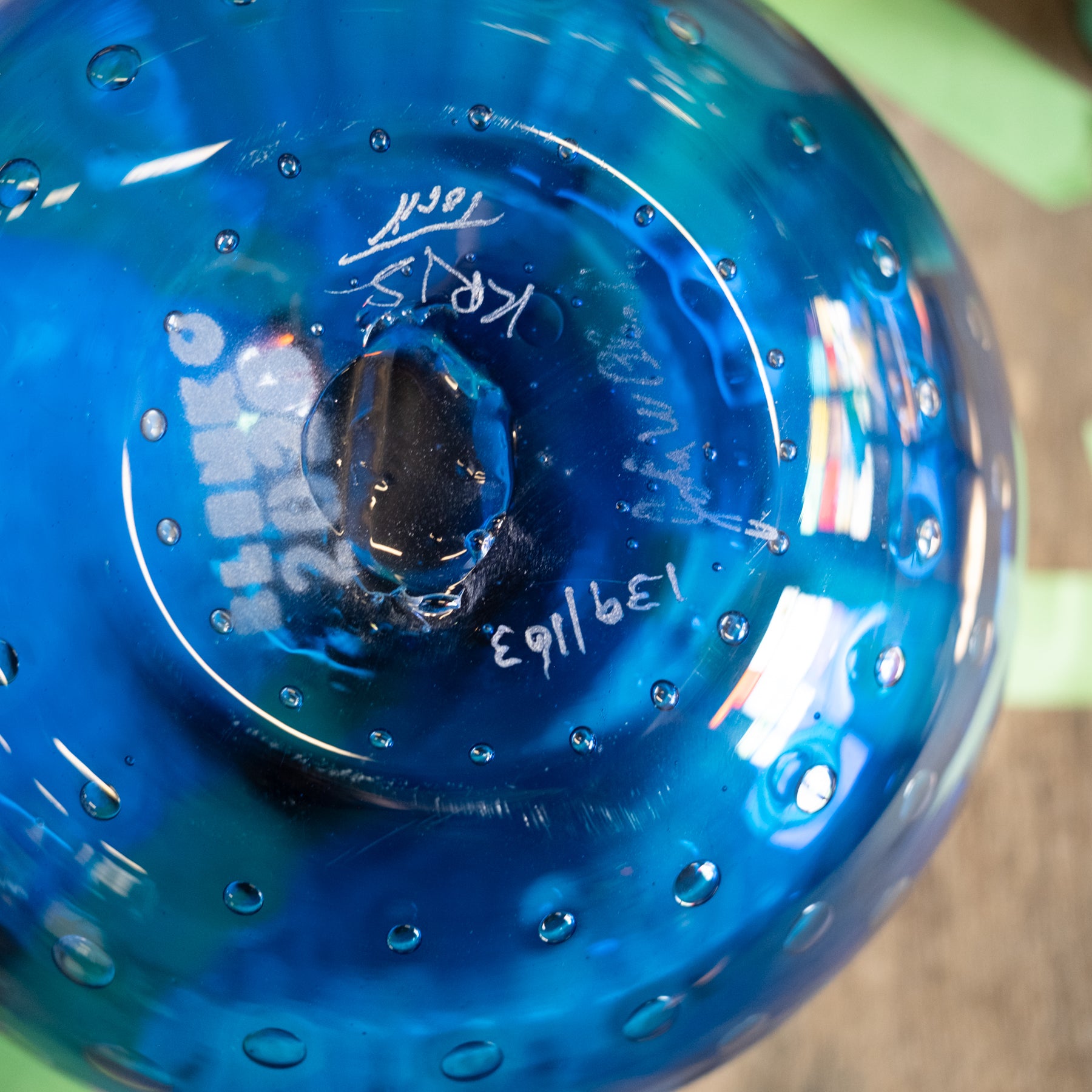 A close-up of a blue glass bowl or vase showing the bottom of the vessel with white hand-written markings and a glossy, bubble-textured surface.
