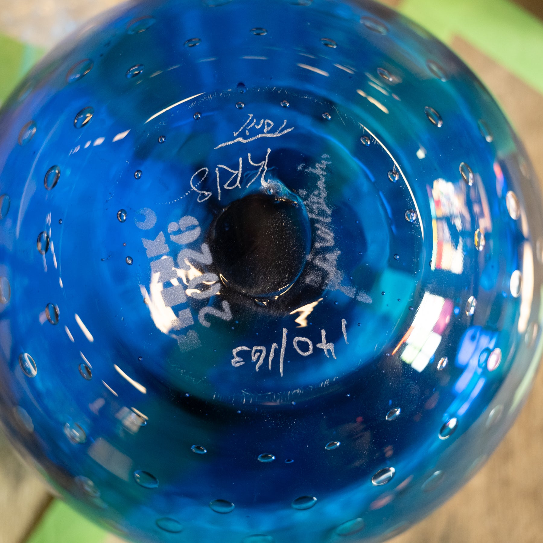 A close-up of a blue glass container or bottle viewed from the bottom, showing a circular opening with small bubble-like perforations around the rim and handwritten white markings on the inside.