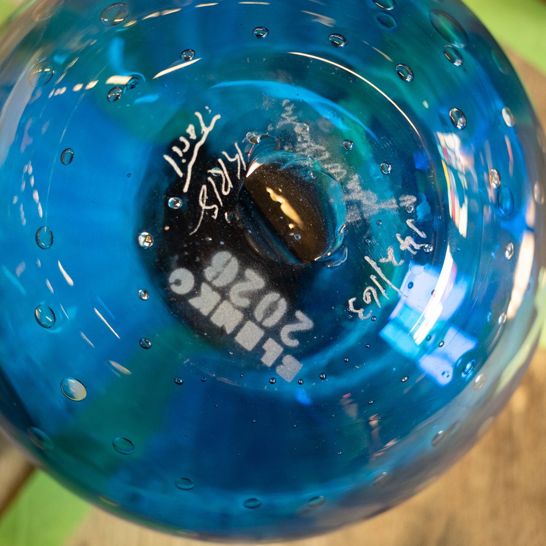 A close-up of a blue glass bowl or vase with a glossy finish, visible air bubbles, and white printed text and signatures on the bottom.
