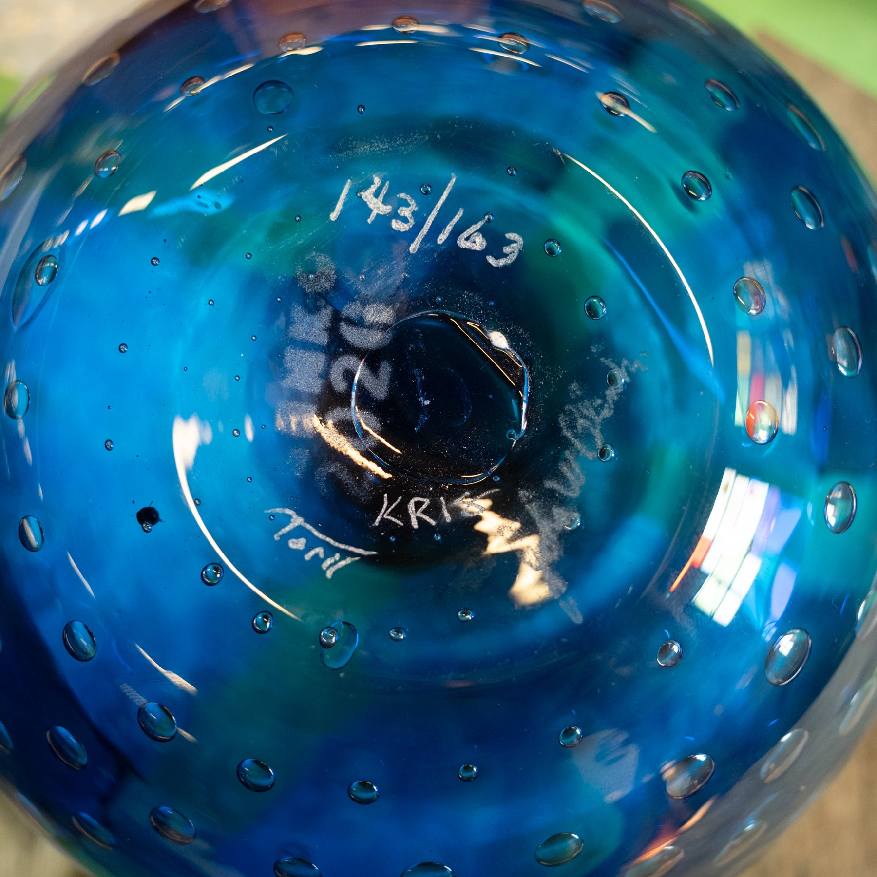 Close-up of the bottom of a blue glass bowl with hand-painted white markings and bubble details, showing the maker's signature 'Kris Torri' and production number '143/163'.