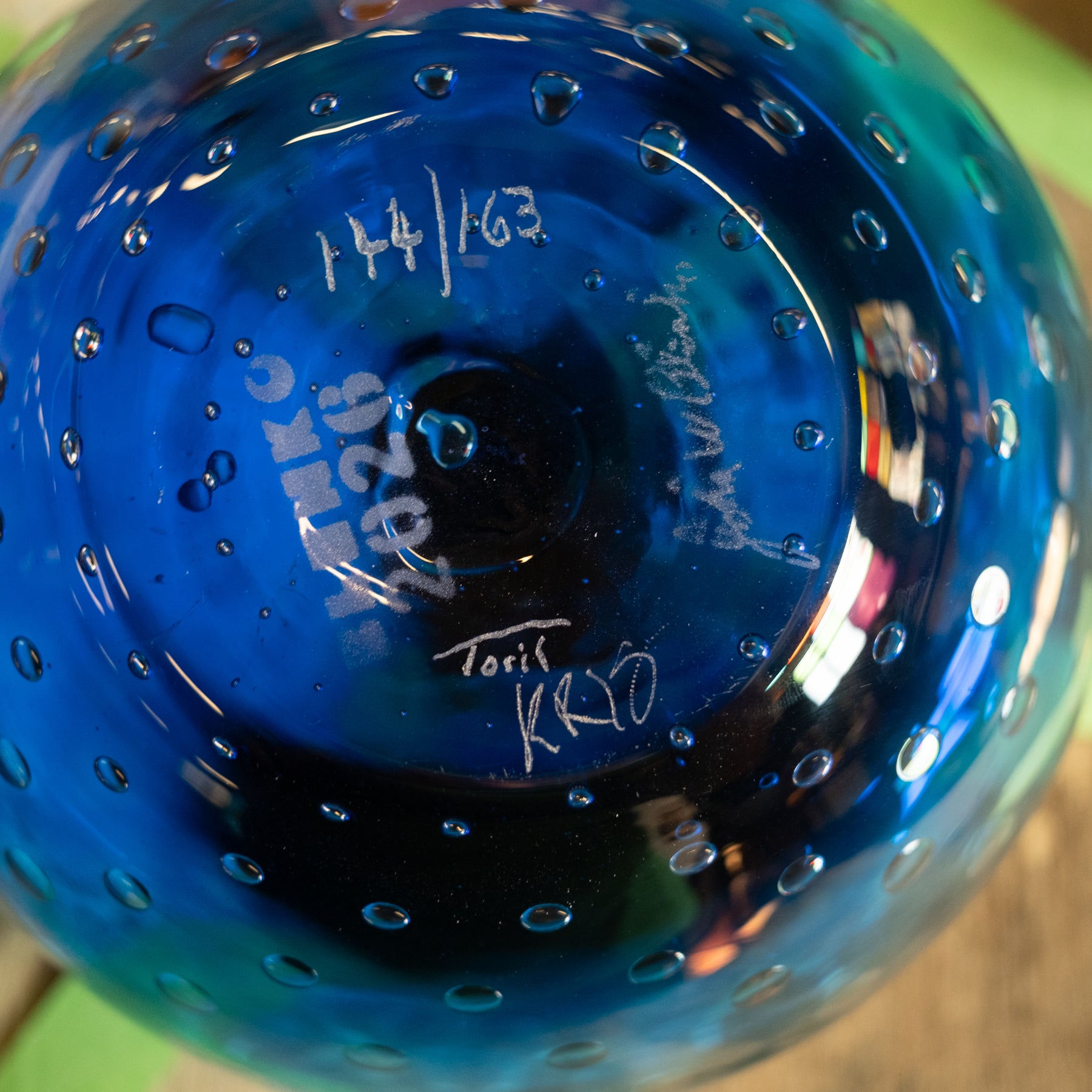Close-up of a blue glass bowl with a perforated bubble pattern, showing white handwritten markings and signatures on the bottom.