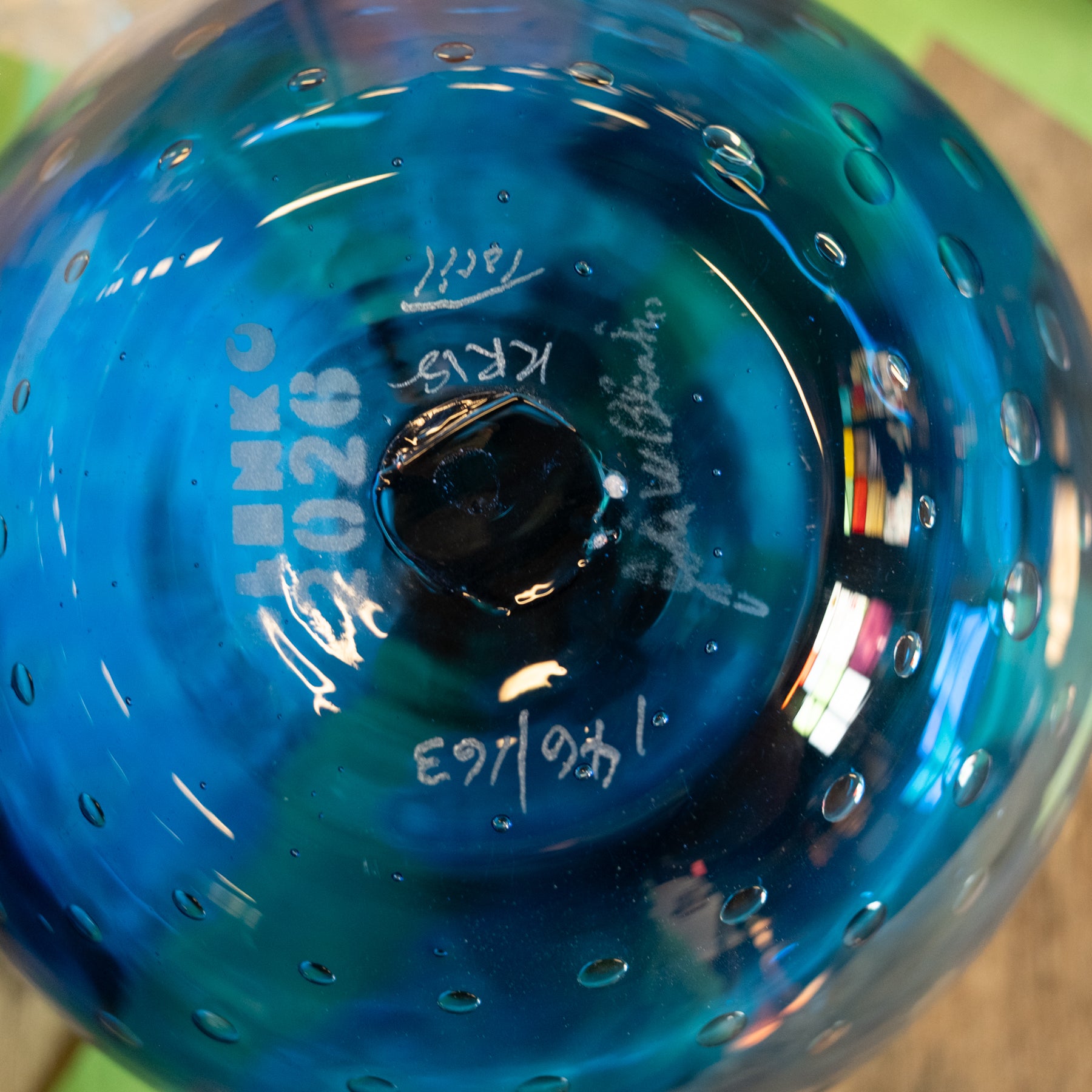 Close-up of the bottom of a blue glass bowl or vase with a circular perforated pattern and white printed markings including 'LENKO 2026' and signatures.