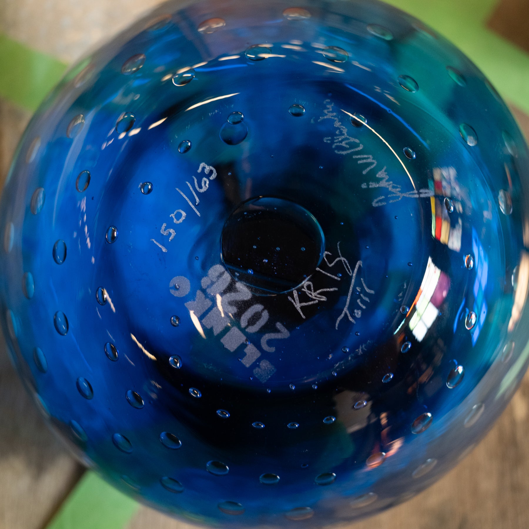 A close-up of the bottom of a cobalt blue glass vase or decorative bowl with a perforated bubble pattern, showing white printed markings and signatures.
