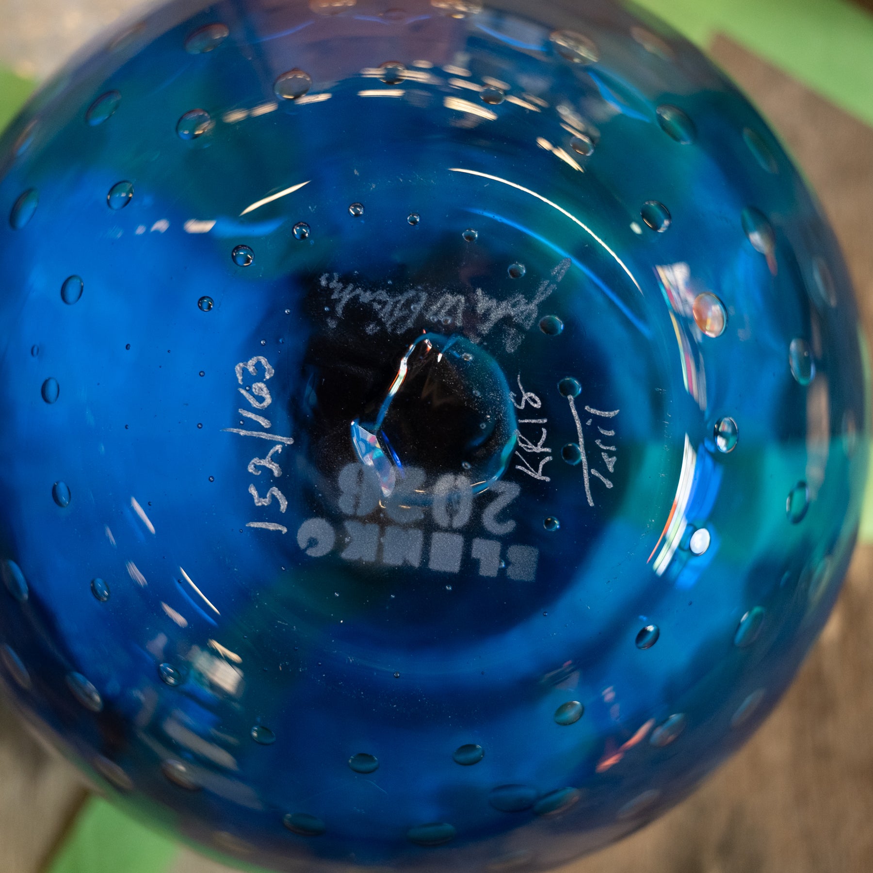 A close-up of a blue glass ornament or vase viewed from the bottom, showing a circular base with bubble-like perforations and white printed markings including 'BLENK' and handwritten dates.