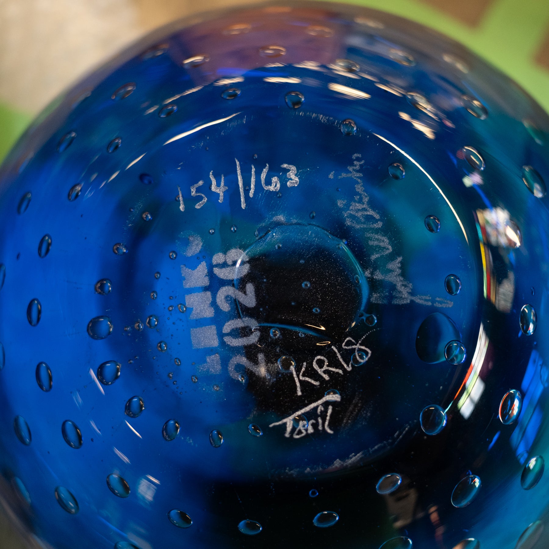 Close-up of the bottom of a blue glass bowl with bubble perforation, showing white handwritten markings including '154/63', '2023', and signatures.