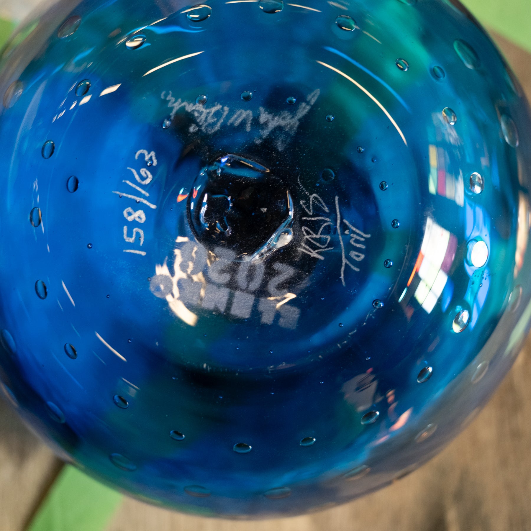 A close-up of a blue glass bowl viewed from above, showing a glossy, bubble-textured surface with white hand-written markings and signatures on the bottom.