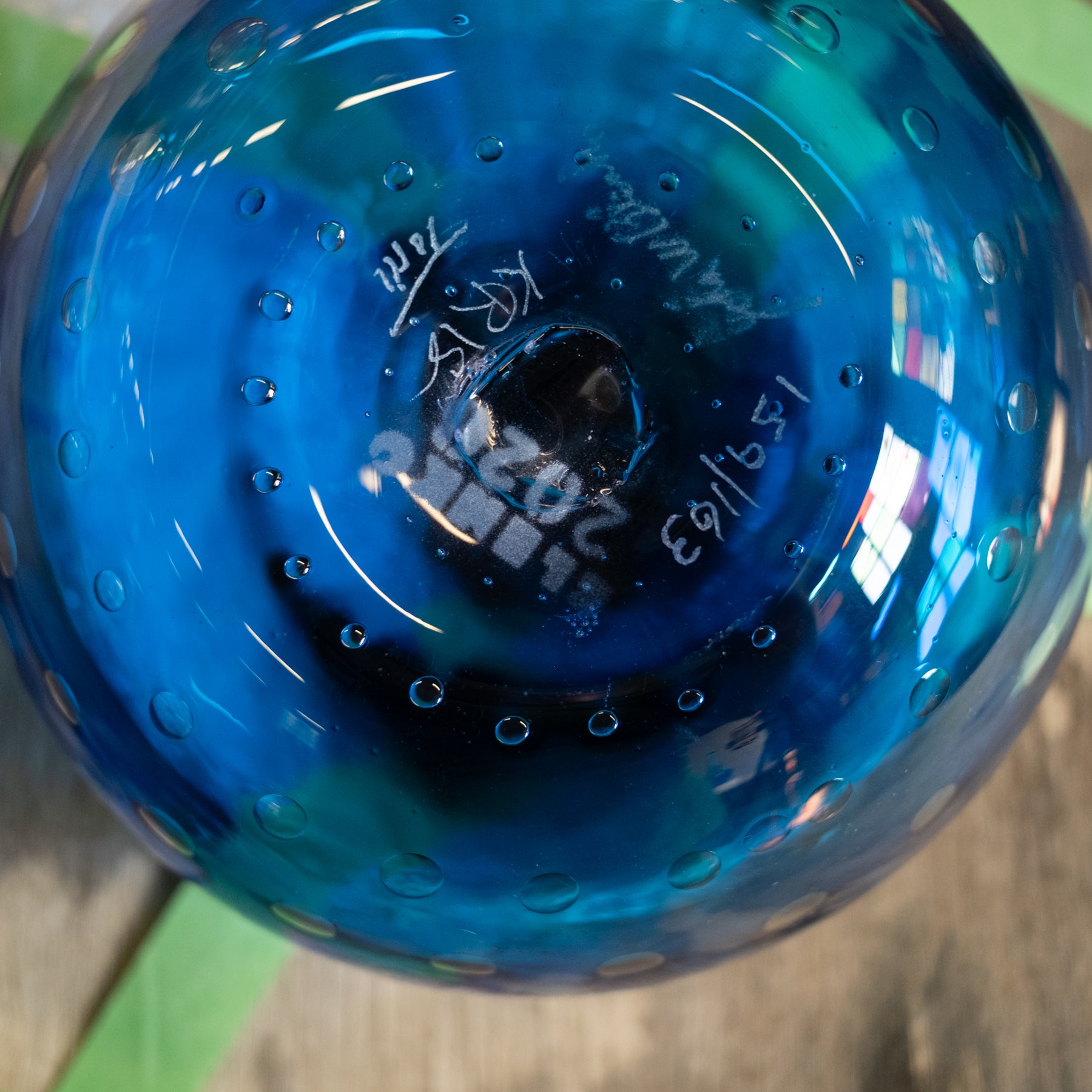 A close-up of a blue glass ornament or vase viewed from the bottom, showing a textured bubbled surface and hand-written maker's marks in white ink.