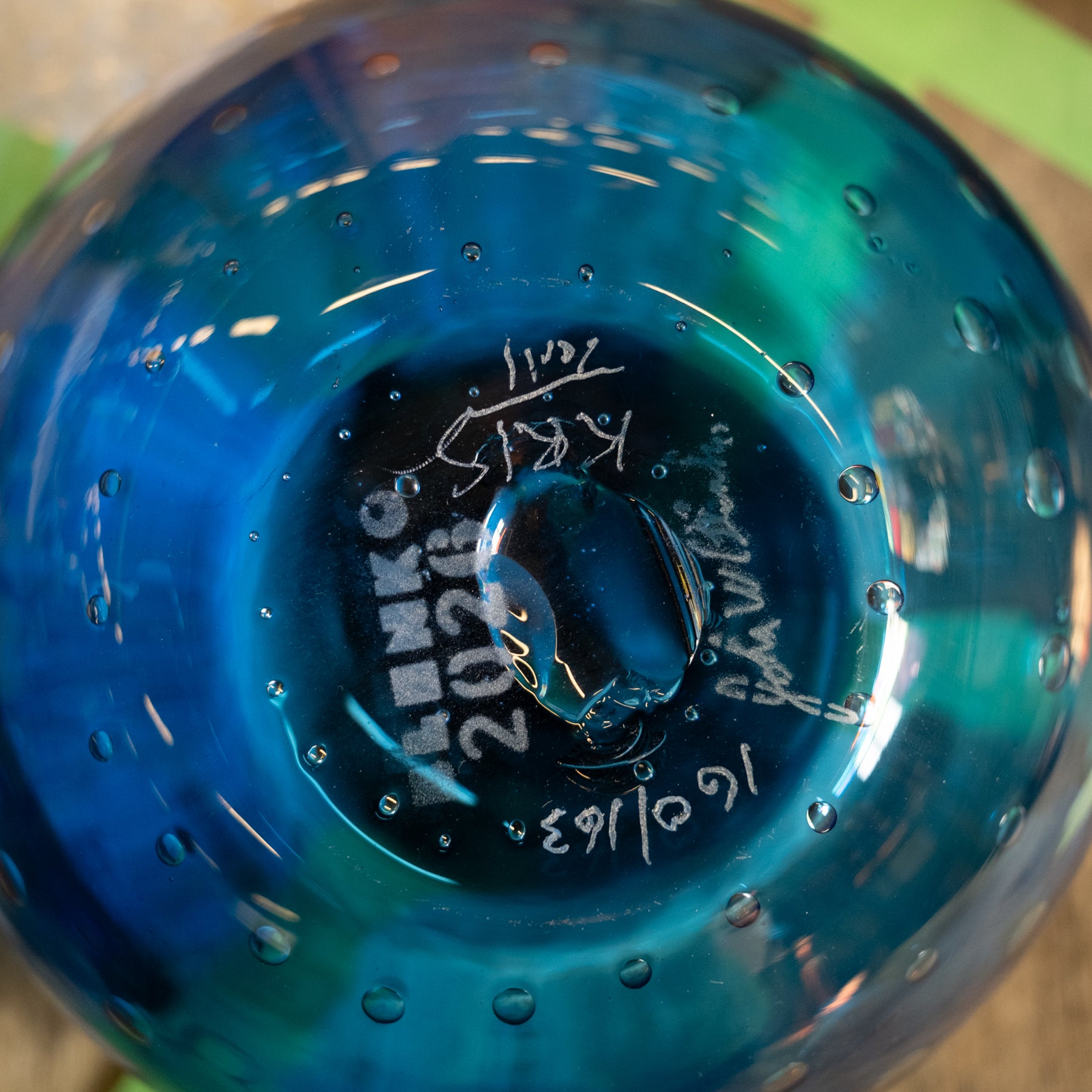 Close-up of the bottom of a blue glass bottle or vase with water droplets on the surface. White handwritten markings are visible on the glass, including the brand name 'Dench' and date '2021'.
