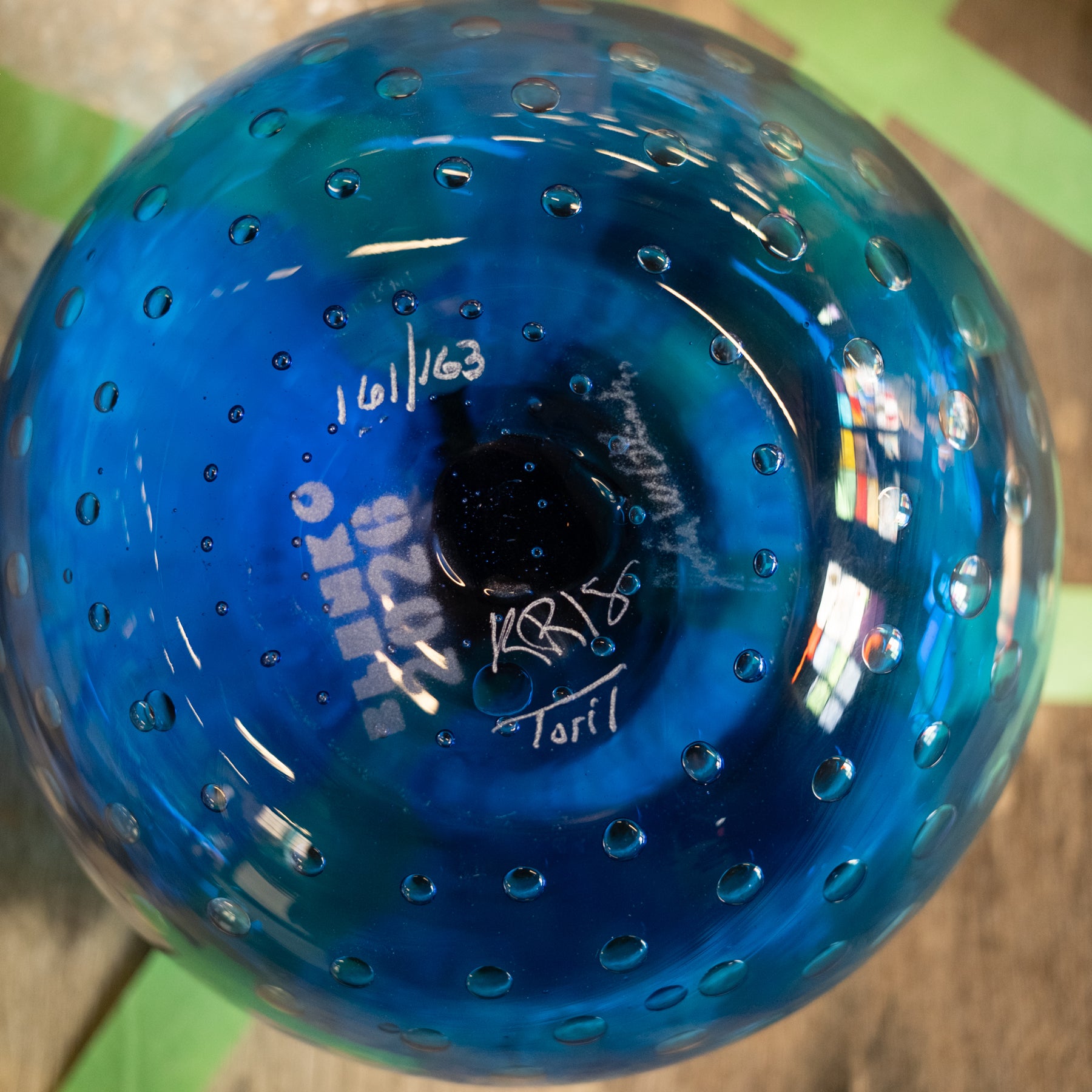 A close-up of a blue glass bowl with a textured bubble pattern and white handwritten markings on the bottom, photographed on a wooden surface with green painter's tape.