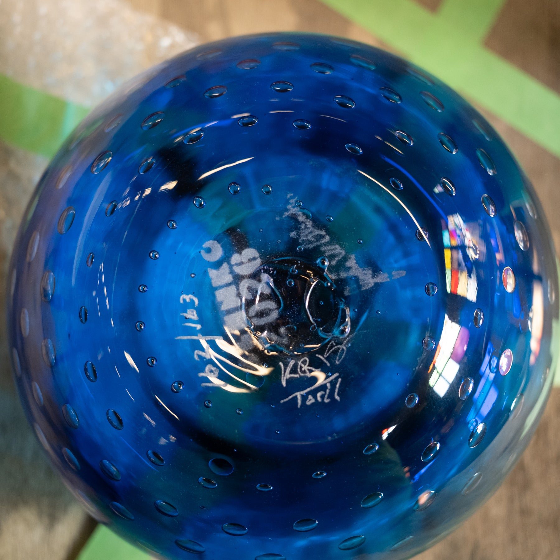 A close-up of a blue glass bowl with a perforated pattern, showing white hand-written markings and signatures on the bottom.