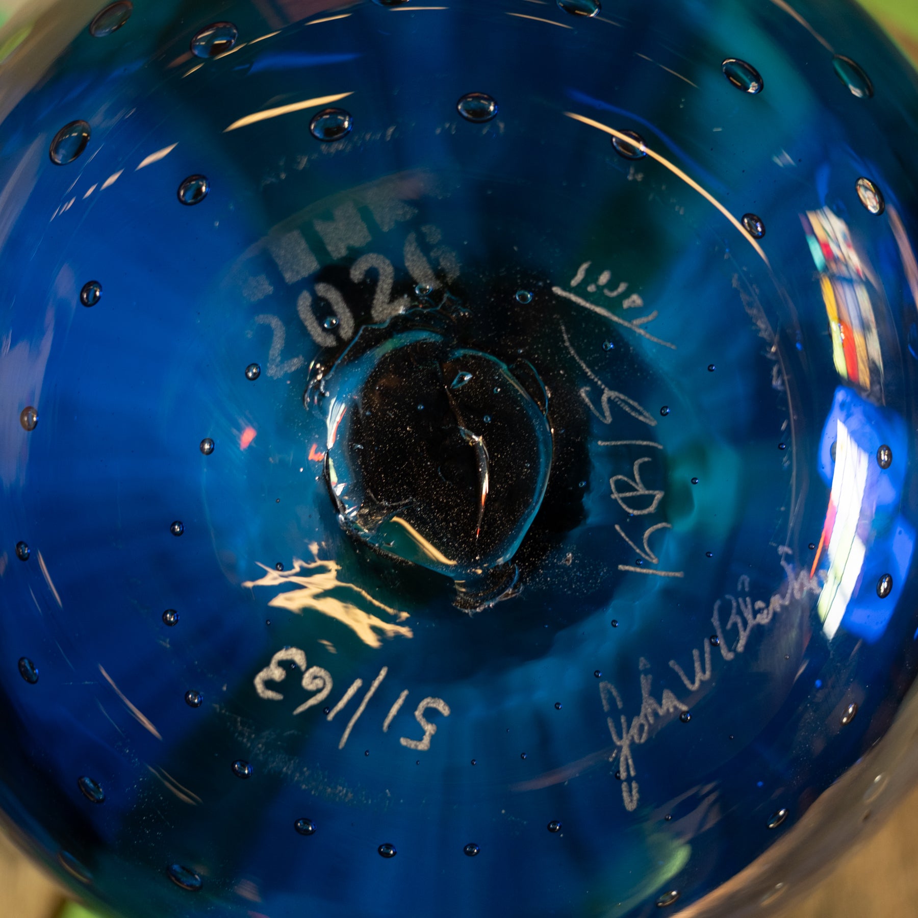 Close-up of a blue glass ornament or decorative bowl with embossed text, signatures, and small bubble details visible on the glossy surface.