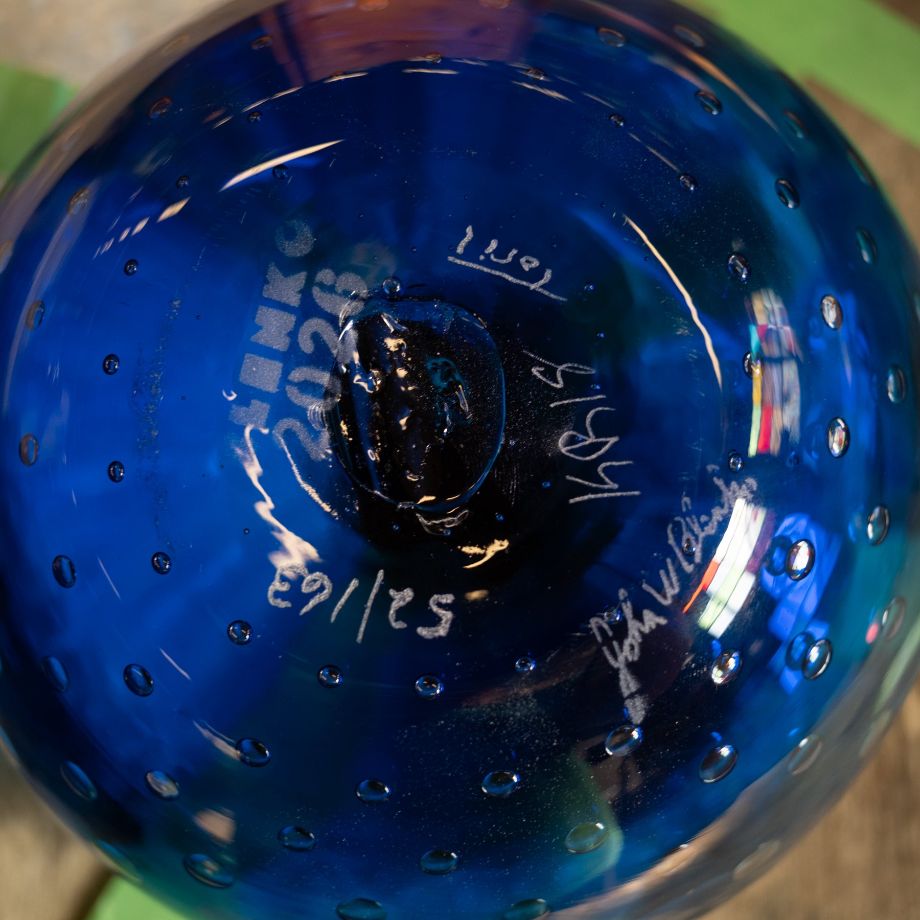 A close-up of a blue glass bowl or vase with a glossy finish, visible white handwritten markings and bubble details on the bottom, and a blurred multicolor background.