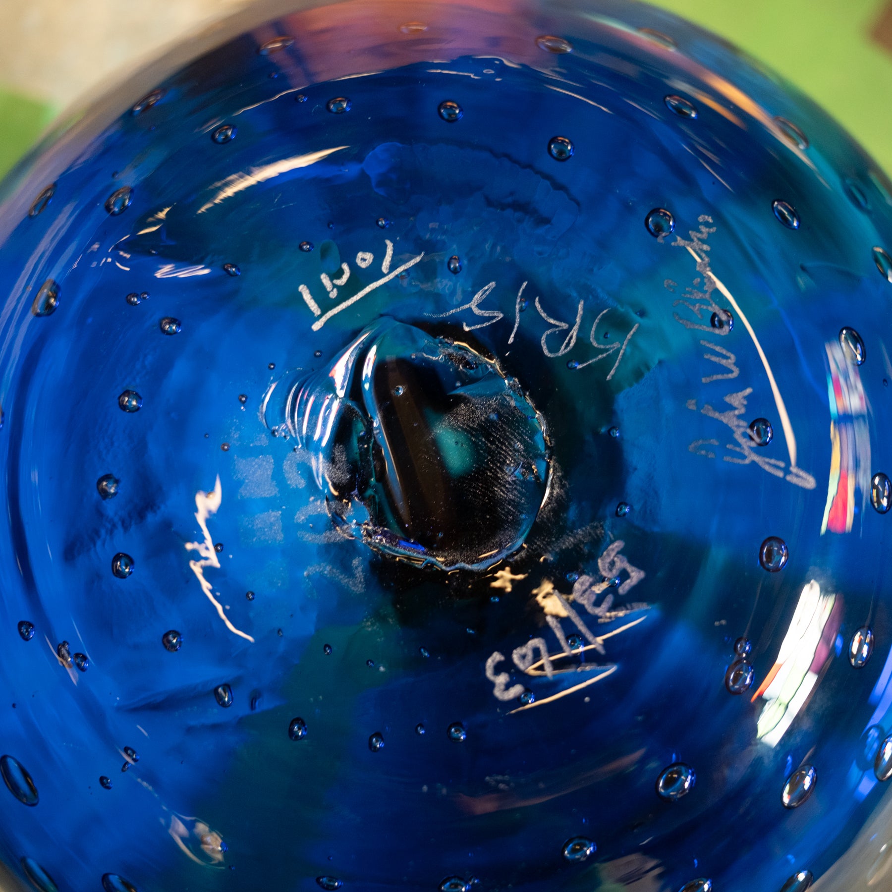 Close-up of the bottom of a hand-blown glass bottle or vase in vibrant cobalt blue, showing the central stem attachment, air bubbles, and white hand-written maker's marks and dates.