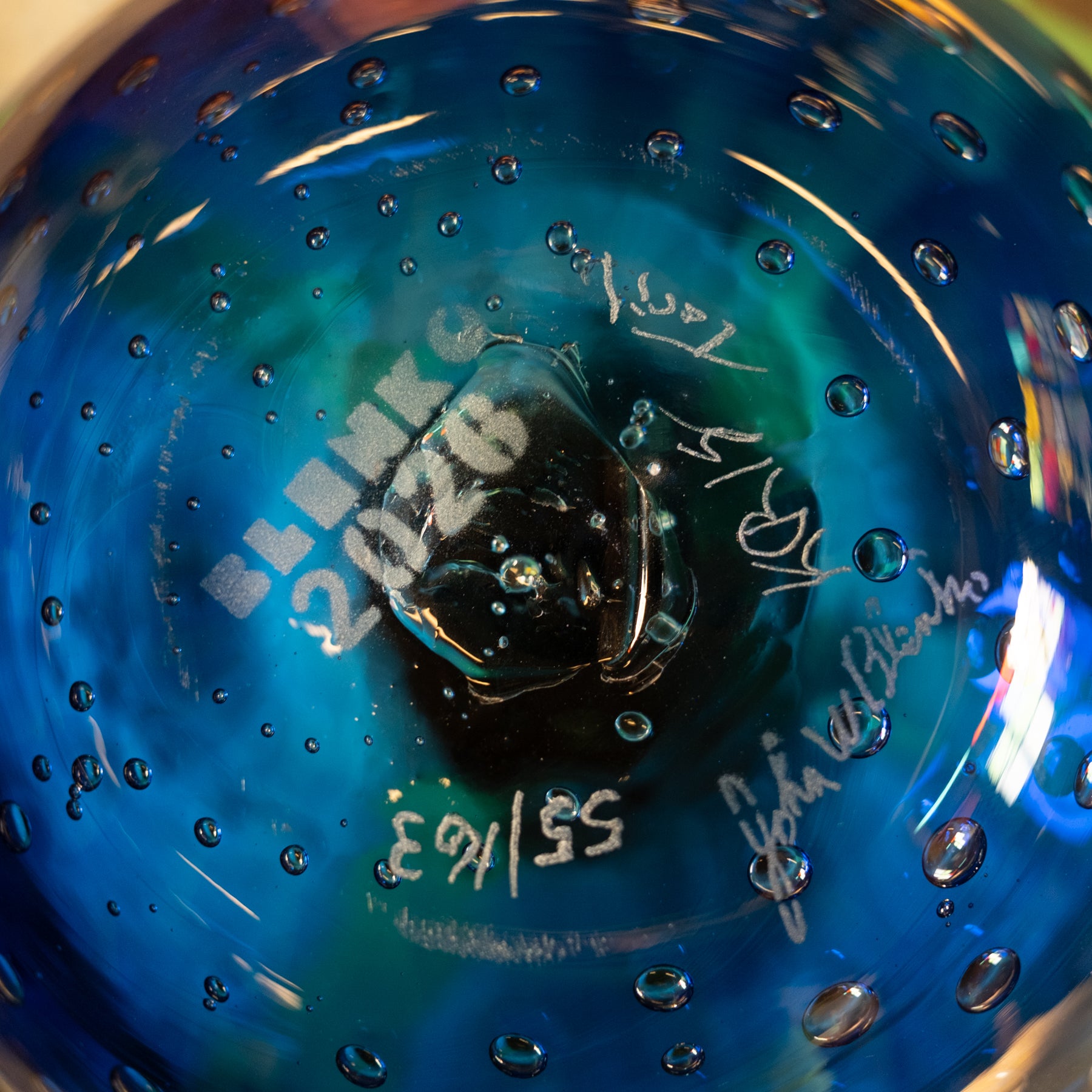 Close-up of a blue glass beaker or flask with visible air bubbles, white printed text reading 'SLINK 2026', and handwritten signatures or markings on the glass.