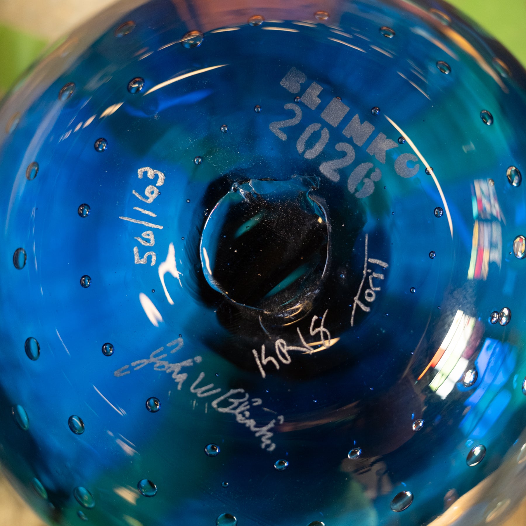 Close-up of the bottom of a blue glass bottle or vase with embossed text reading 'BLUMK 2026', handwritten signatures, and small raised dots around the surface.