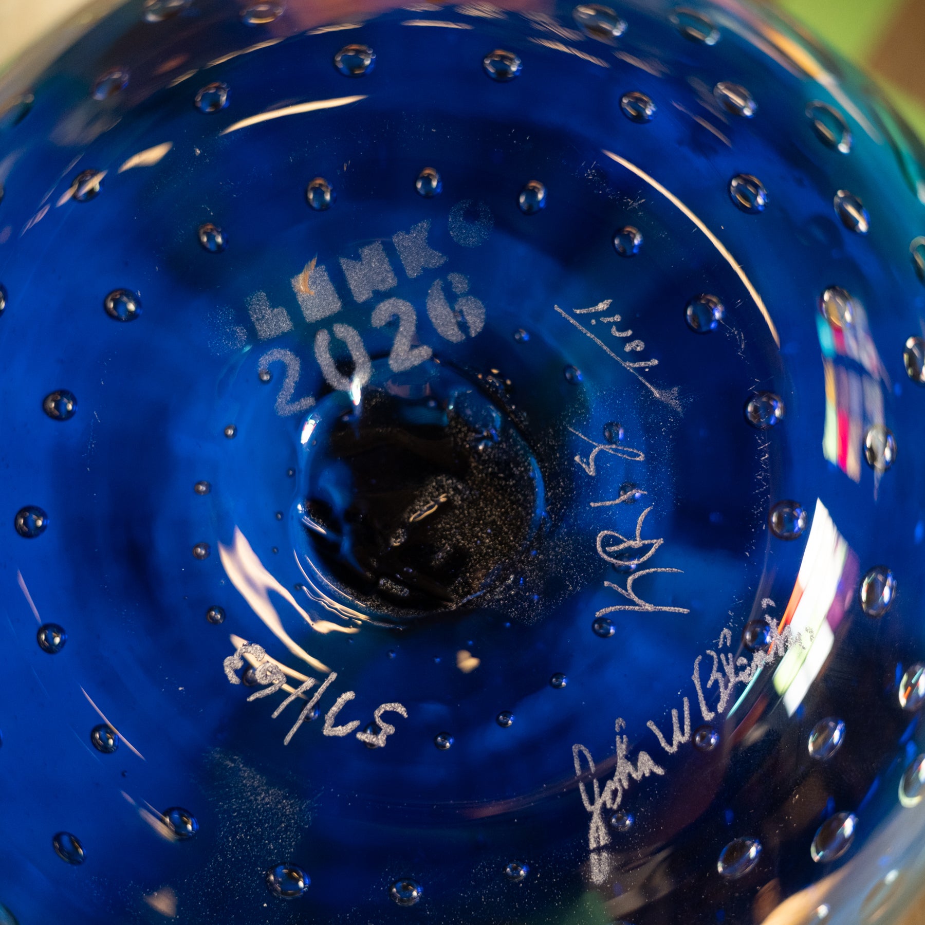 Close-up of the bottom of a cobalt blue glass bowl with a textured bubble pattern, featuring white printed text reading 'BLINKS 2026' and several signatures.