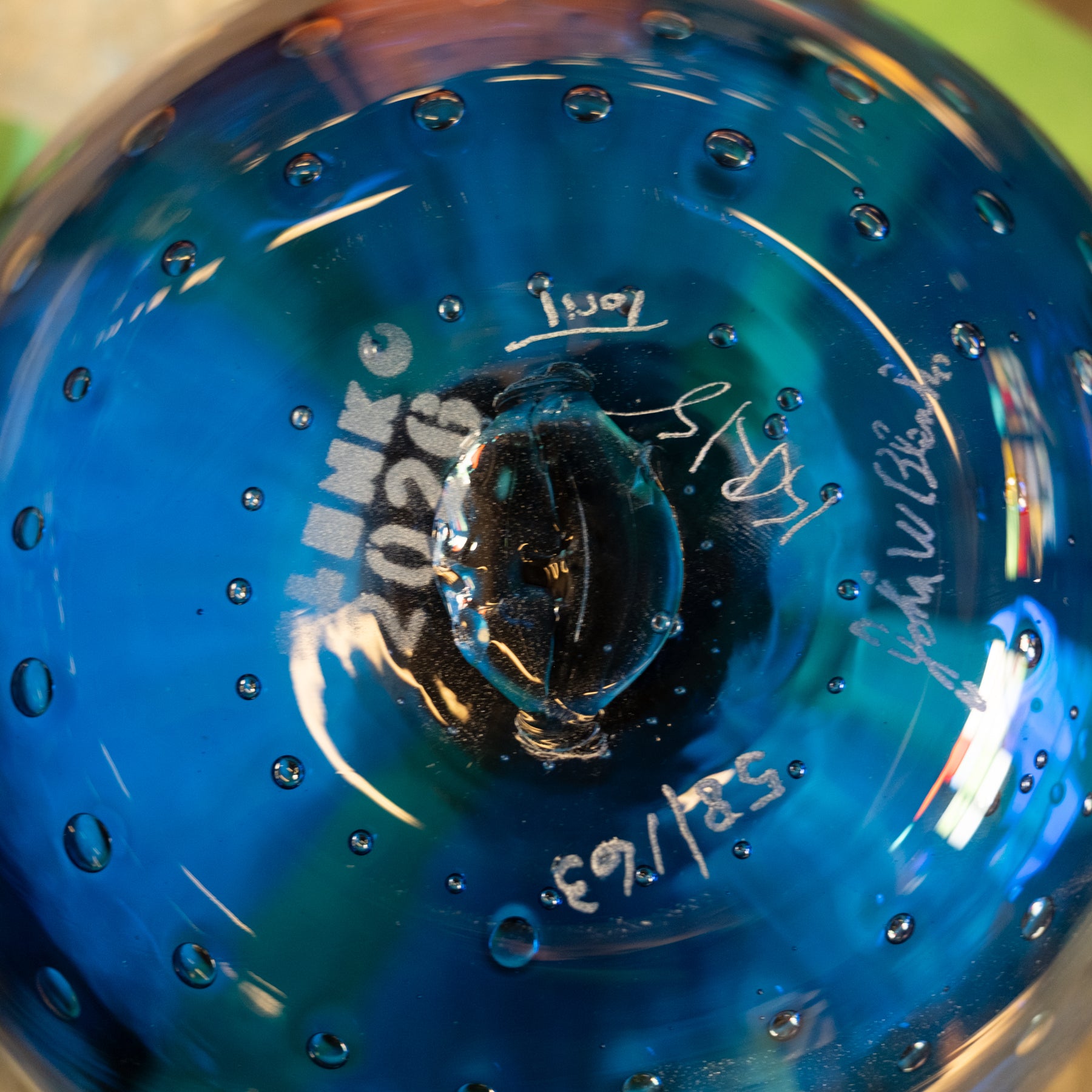 Close-up of a blue glass water bottle base with printed text 'BENKO 2026' and white handwritten markings, showing water droplets on the glossy surface.