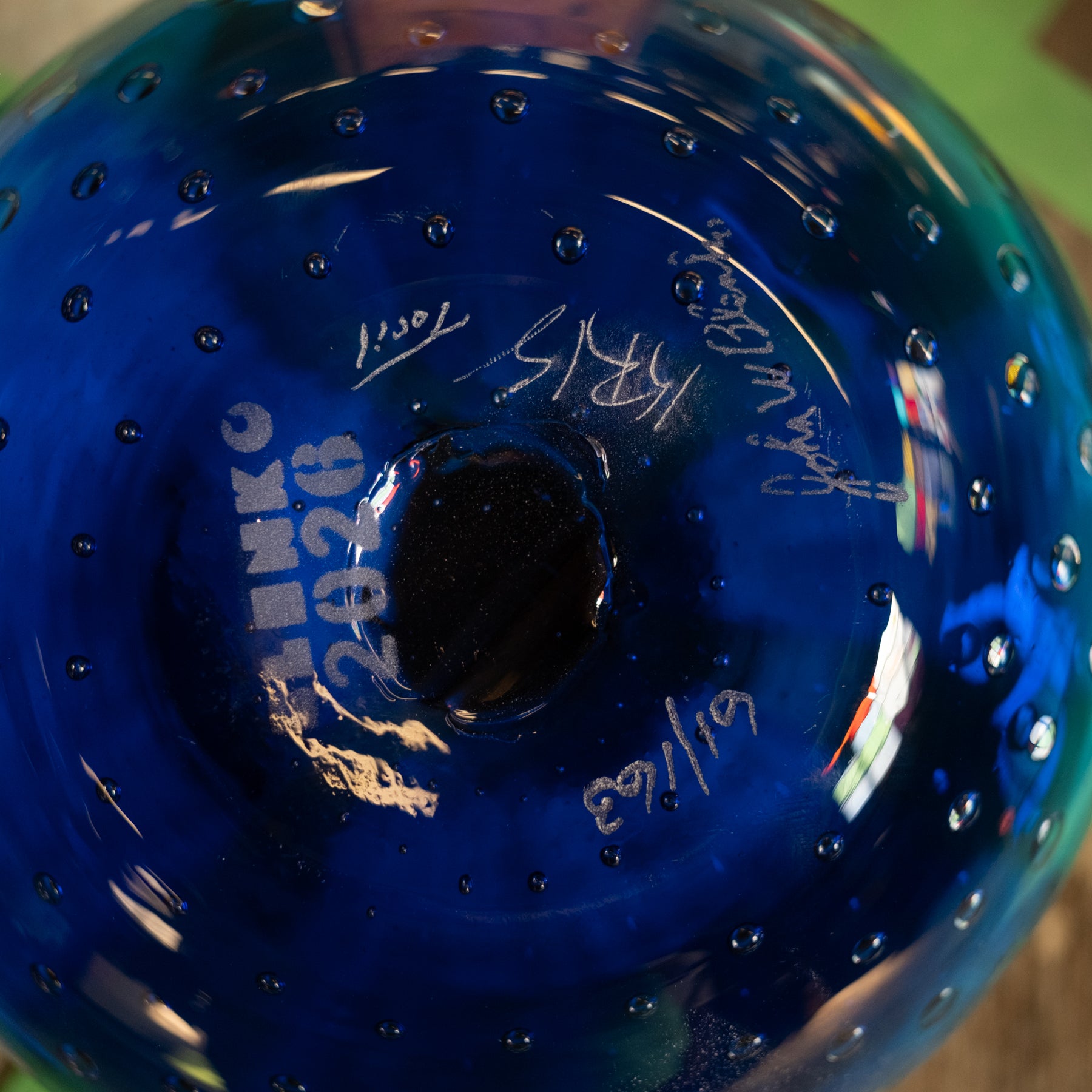 Close-up of the bottom of a blue glass bowl or vase with a circular hole in the center, showing white printed text and signatures on the glossy glass.