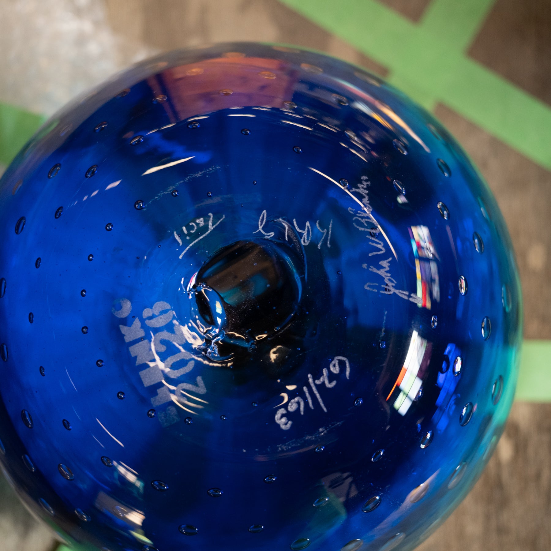 A close-up of a glossy cobalt blue glass bowl with a perforated rim and white handwritten markings on the inside, photographed against a blurred background with green tape.
