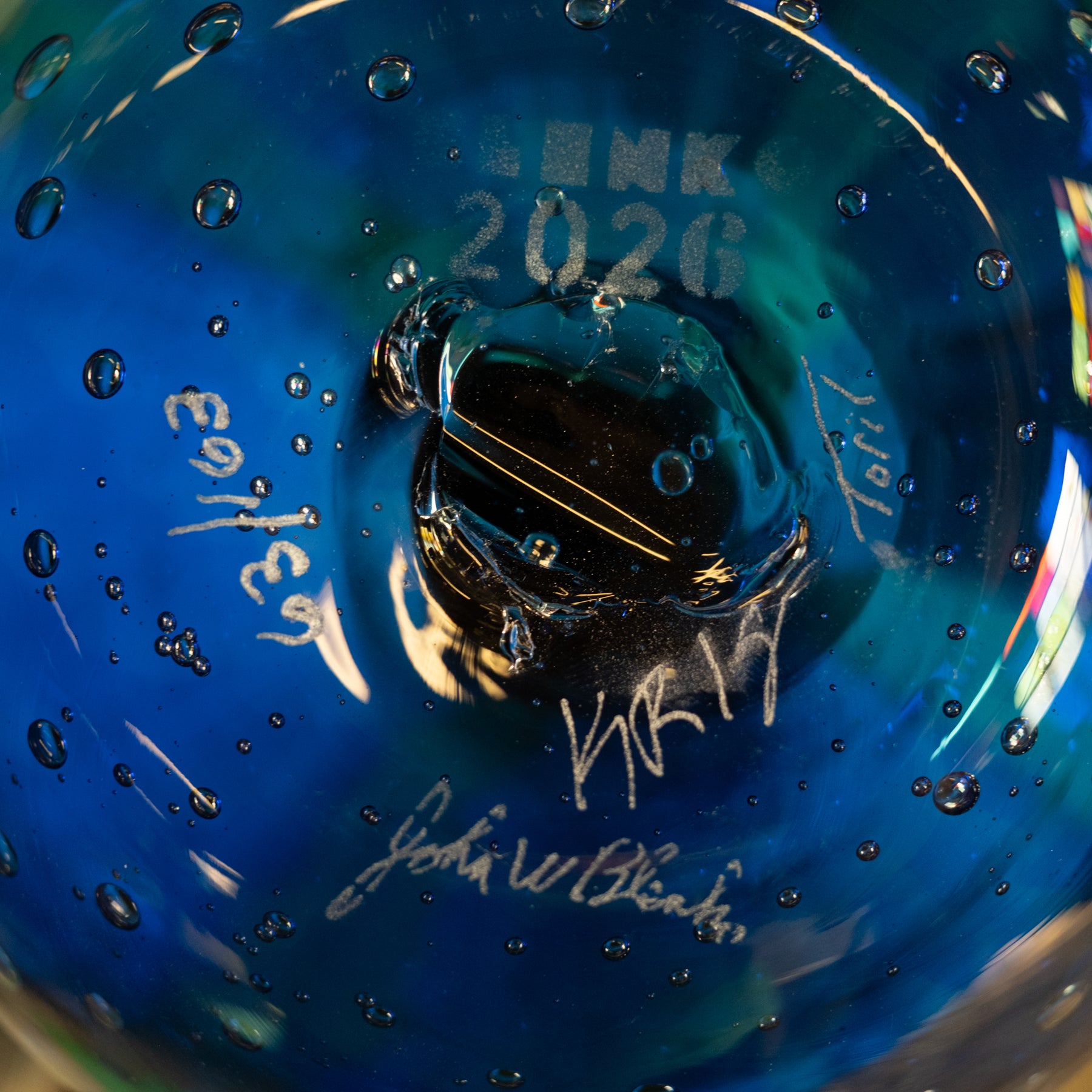 Close-up of a blue glass bowl or vase with water droplets, featuring etched text reading 'LINK 2026' and several white handwritten signatures.