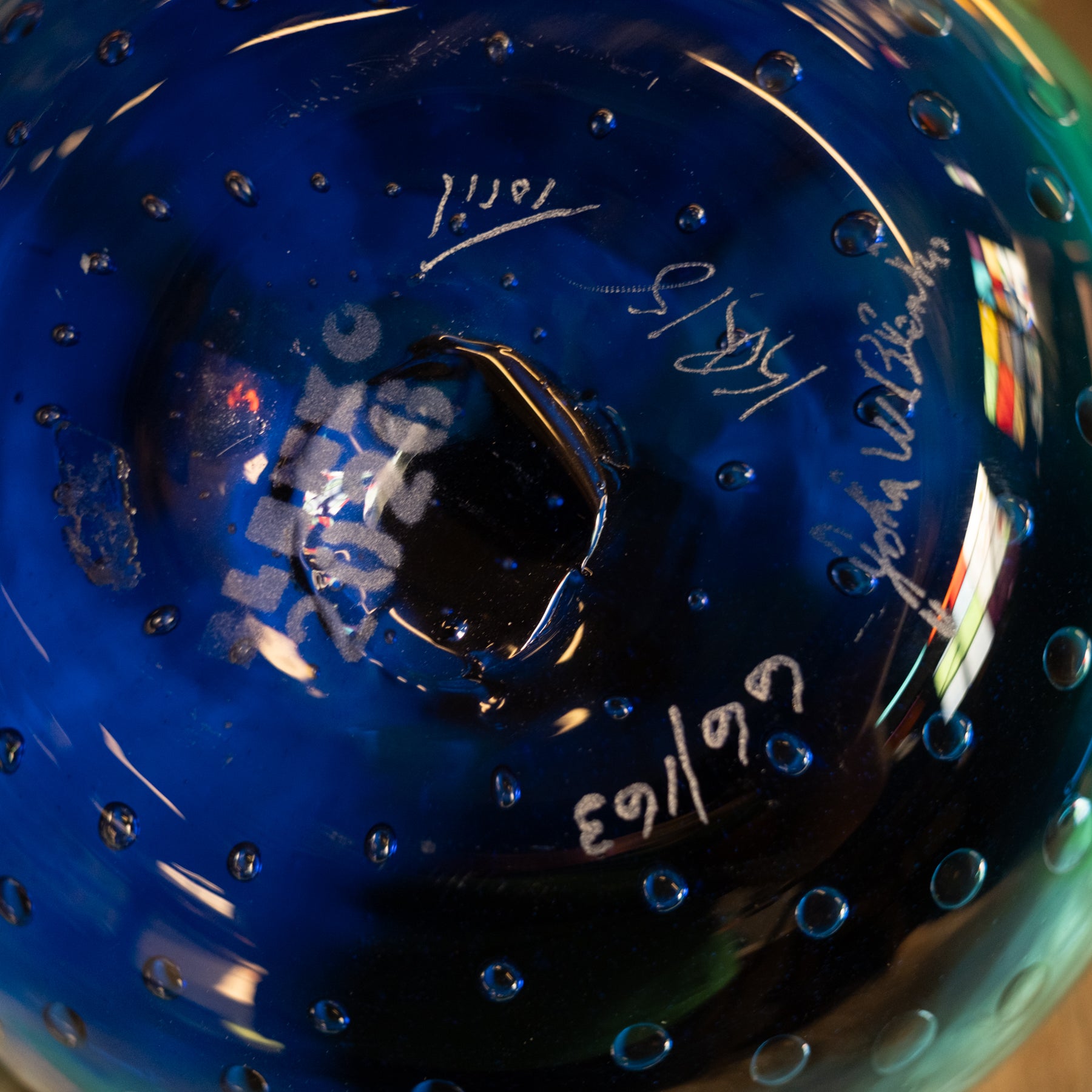 Close-up of a blue glass bowl or vase with a central hole, covered in small raised bubbles and signed with white marker text and dates.