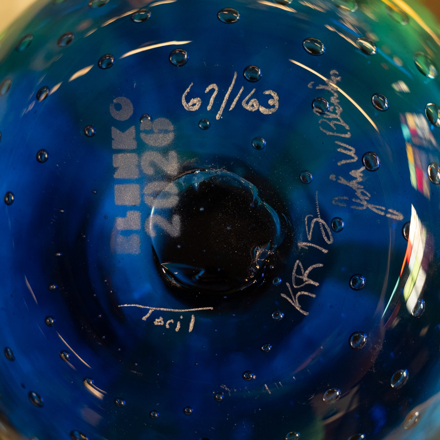 Close-up of a blue glass bowl or vase with a circular center, showing white handwritten markings and signatures on the inside surface.