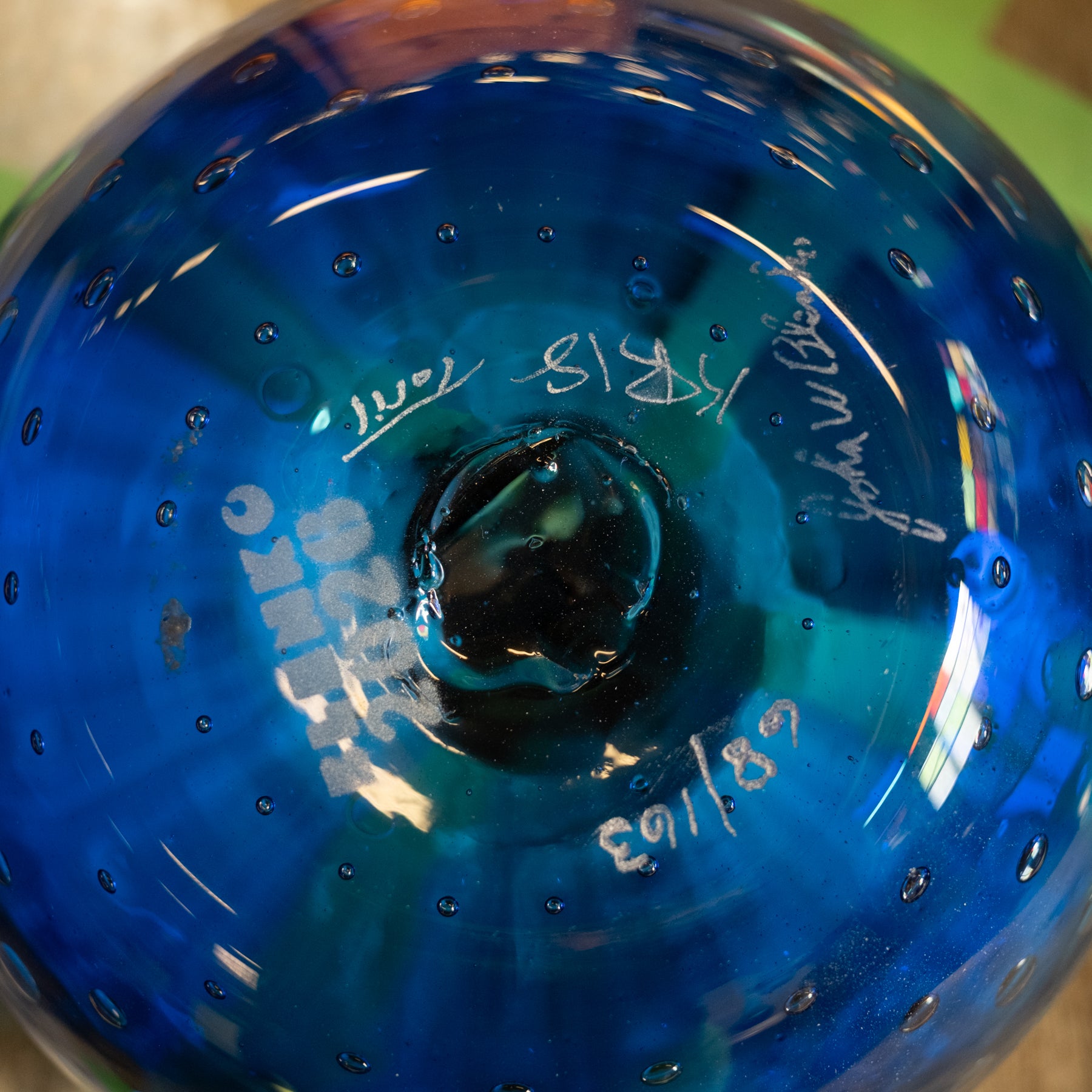 Close-up of a blue glass bowl or vase with a glossy finish, featuring white handwritten markings and bubbles on the surface.