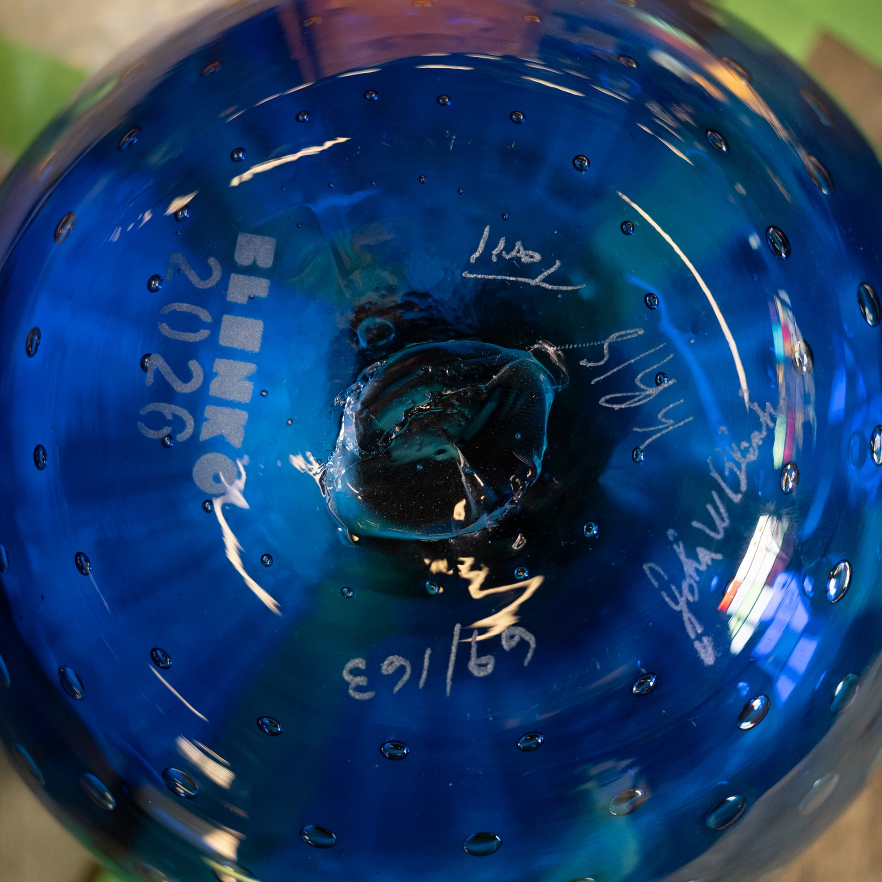 Close-up of the bottom of a blue glass decorative object, showing the brand name 'BLENKROE' printed vertically, the year '2026', and several white handwritten signatures or markings.