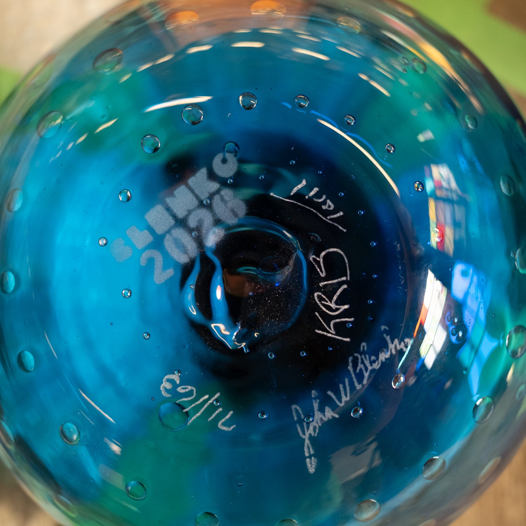 Close-up of a blue glass bowl or vase with a glossy finish, bubble details, and white handwritten signatures and text on the base.