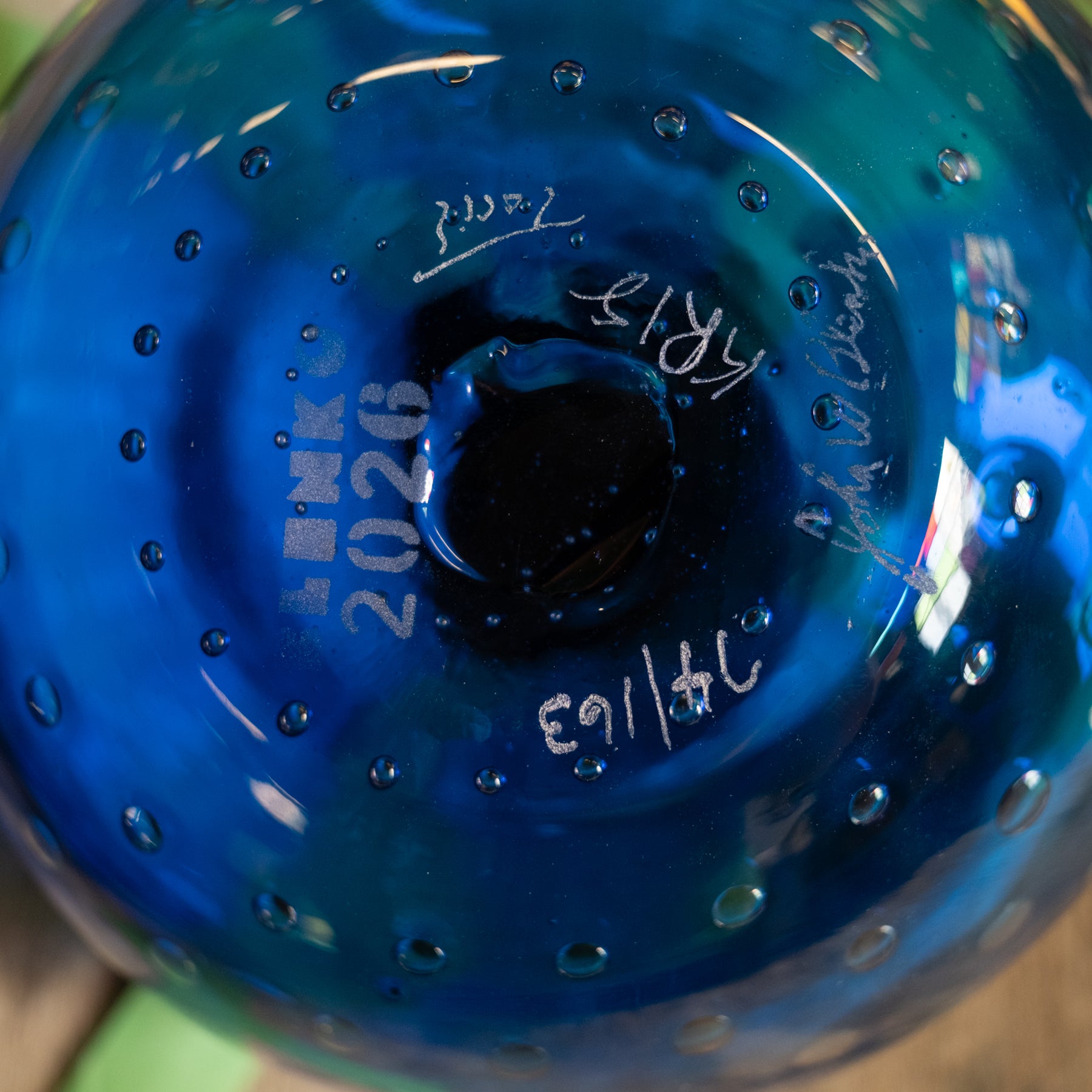 Close-up of the bottom of a blue glass vessel with etched markings including 'LINKS 2026', handwritten dates, and a circular bubble pattern.
