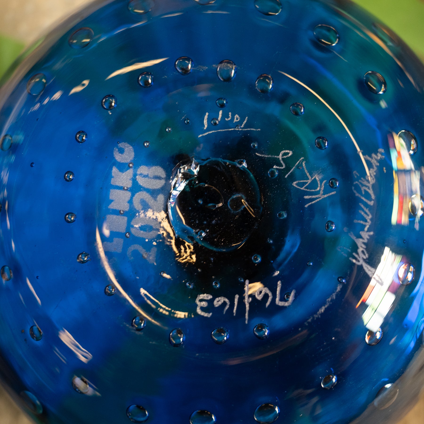 Close-up of the bottom of a blue glass bowl or vase with a textured bubble pattern, featuring white Hebrew text and signatures.