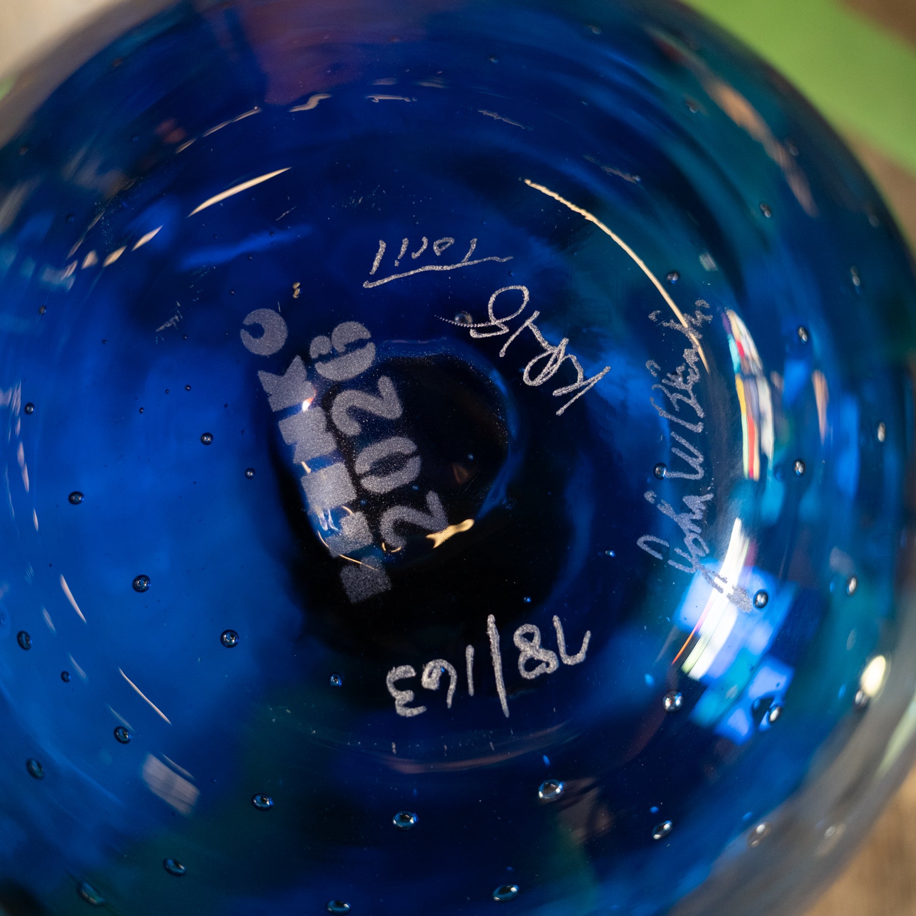 A close-up of the bottom of a vibrant blue glass bowl, showing hand-written white markings and serial numbers.