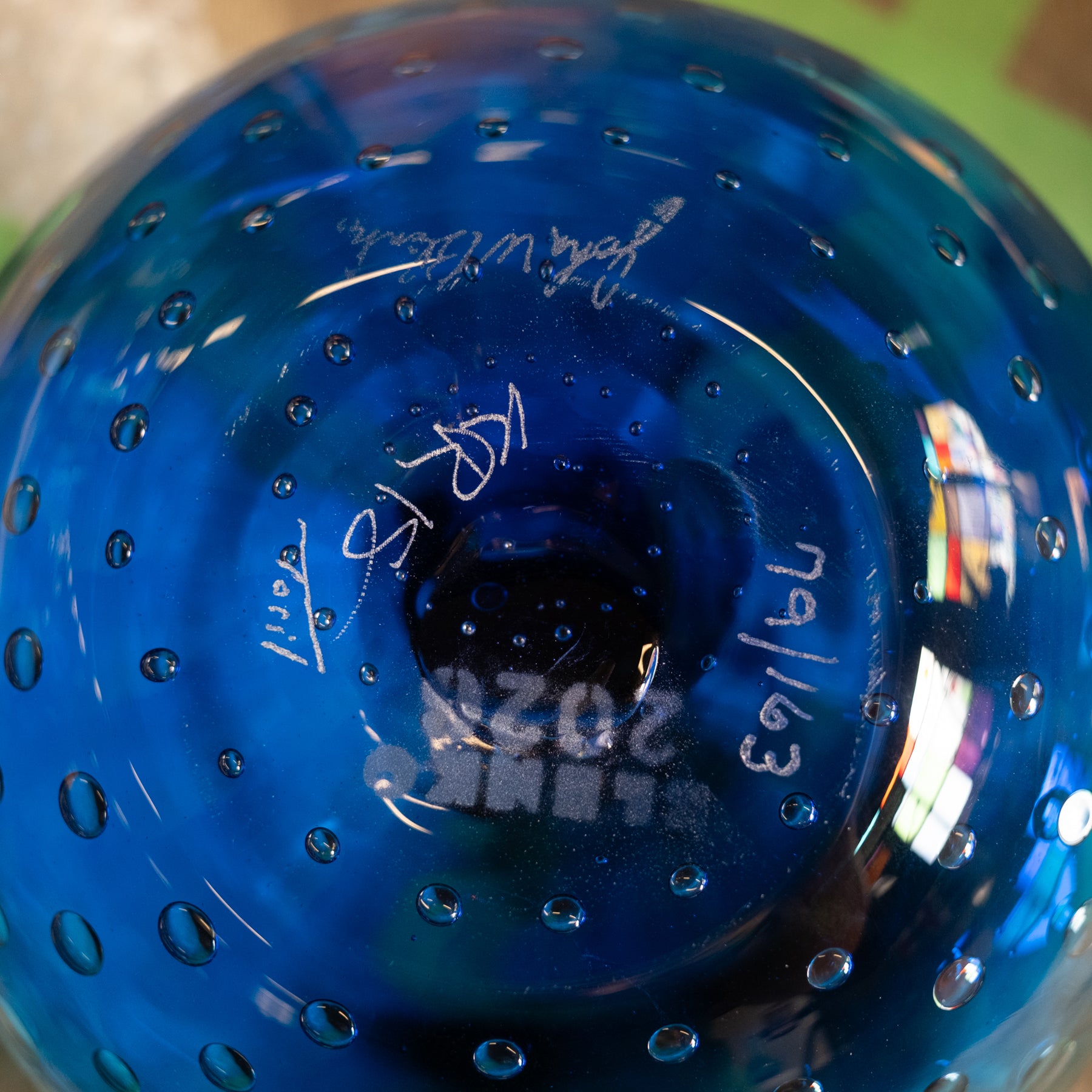 Close-up of the bottom of a blue glass bowl or vase with a perforated bubble pattern, showing white handwritten markings and reflections.