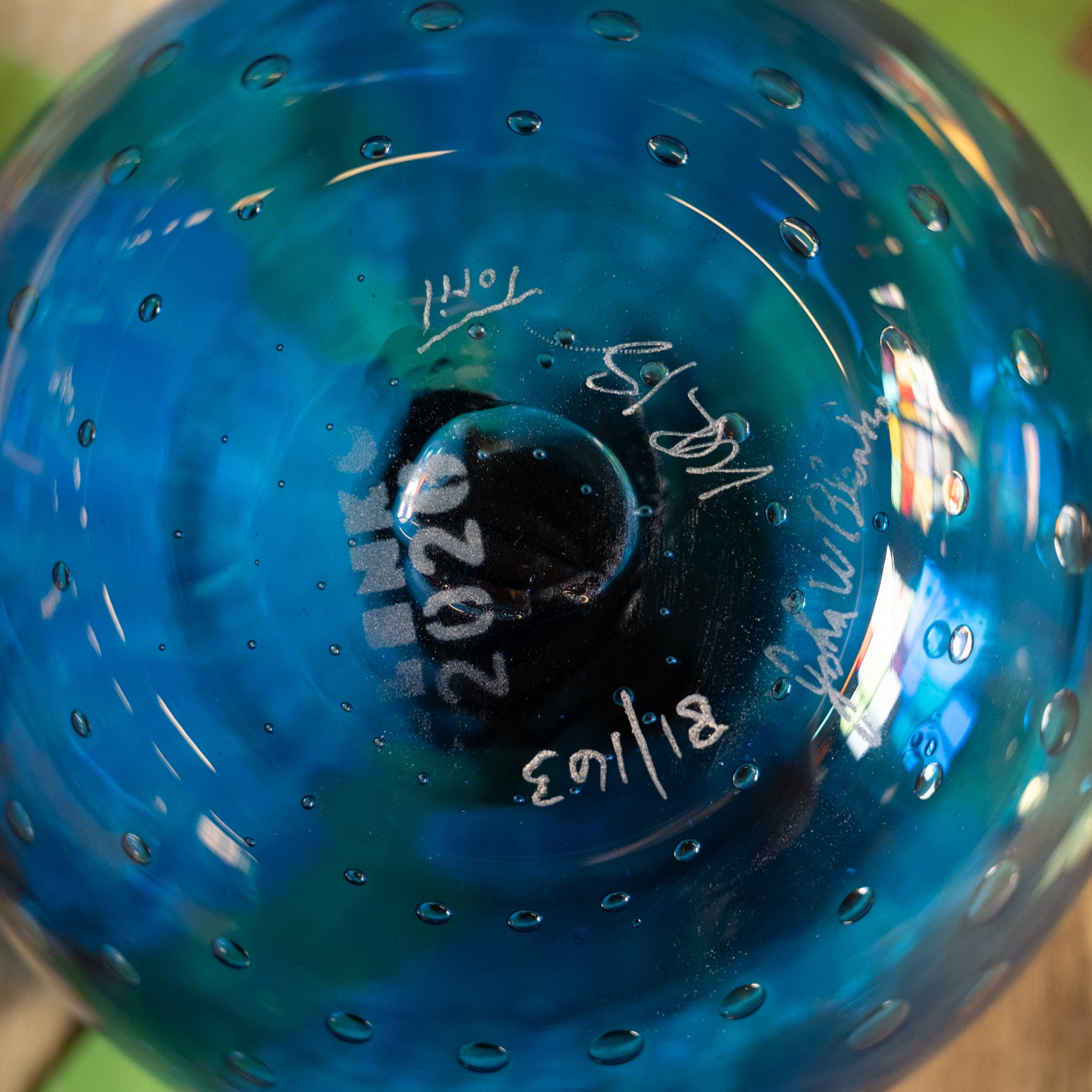 A close-up of a blue glass ornament or vase with a textured bubble pattern and white handwritten markings on the bottom.