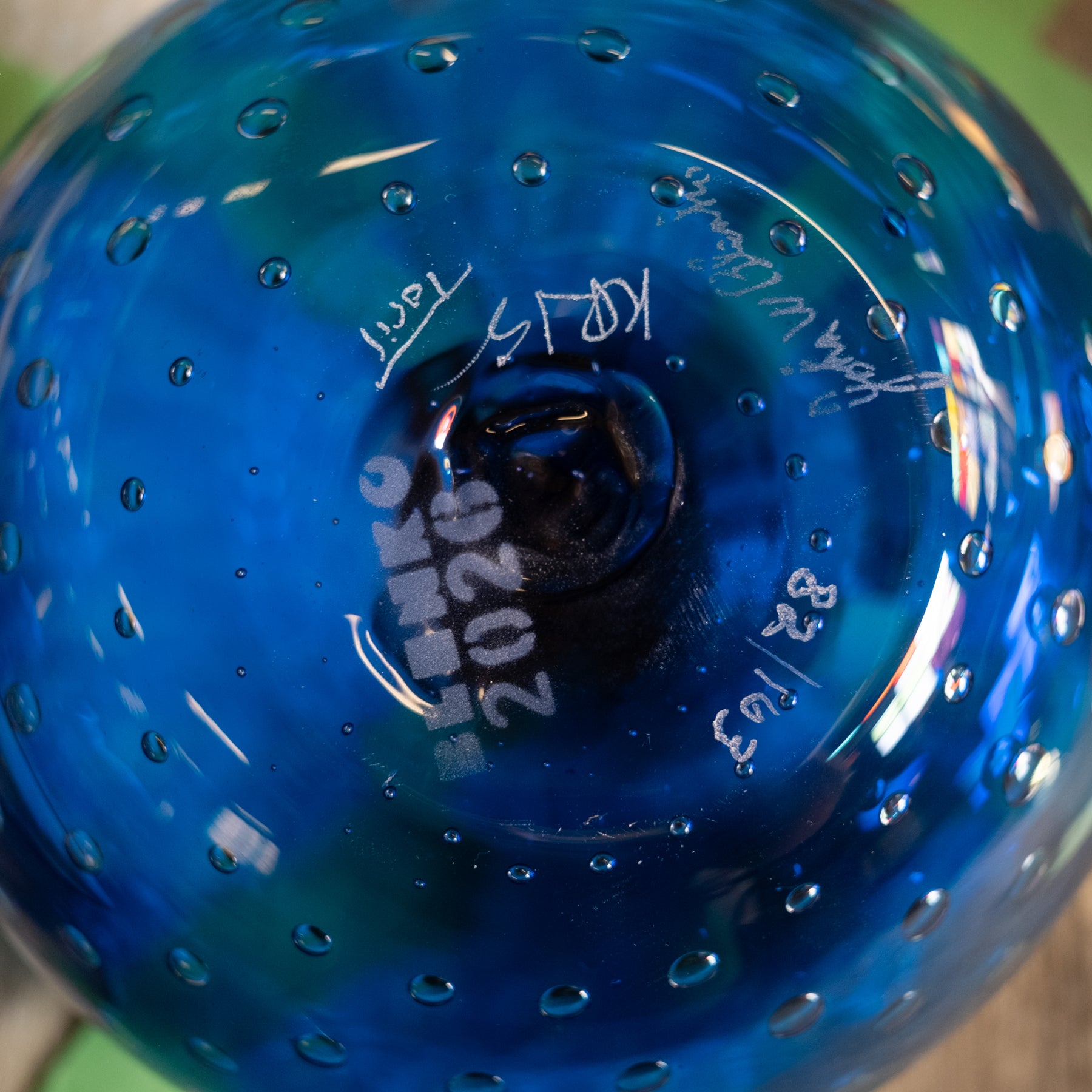 Close-up of a blue glass object, likely a decorative bowl or vase, showing embossed white text and signatures on the bottom surface.