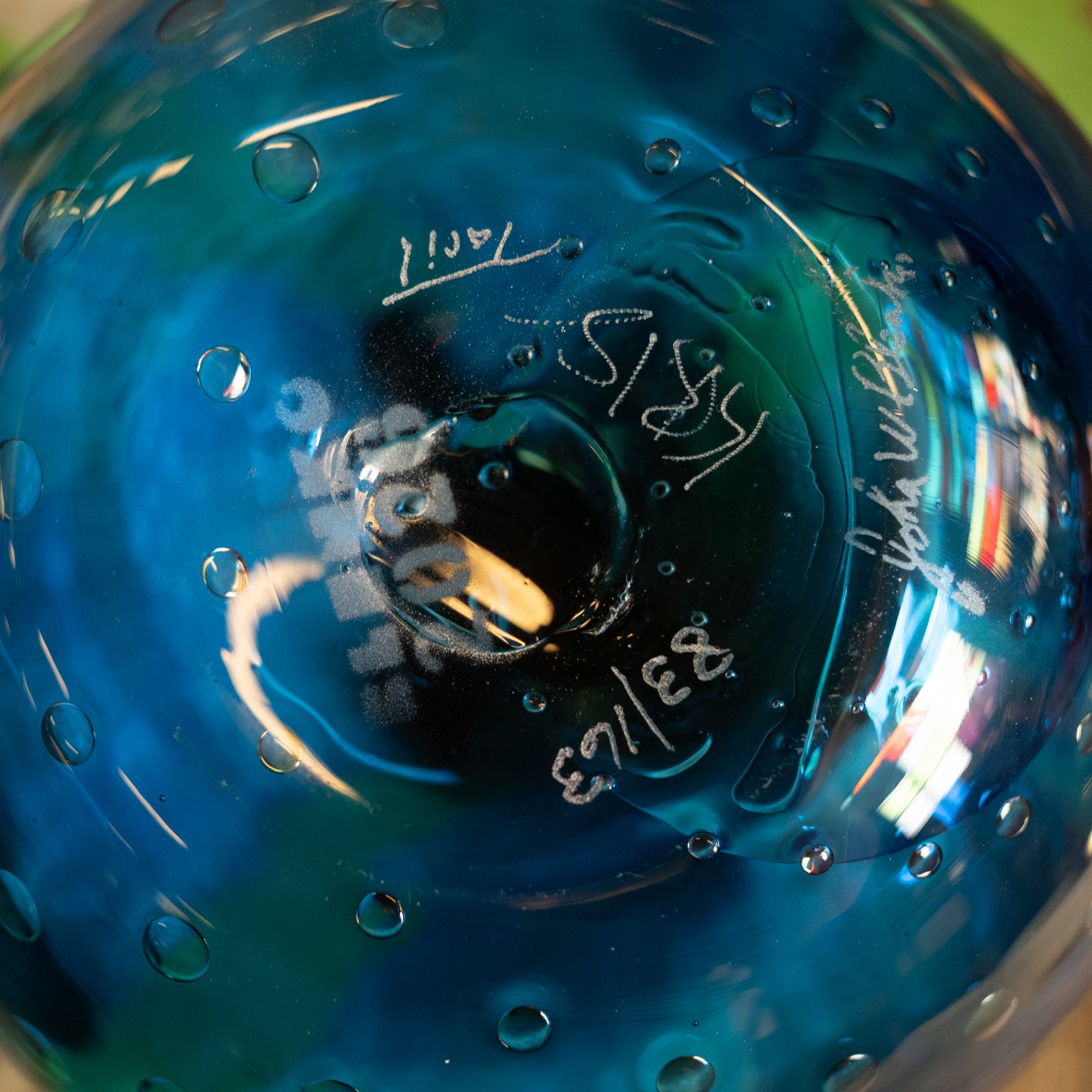 Close-up of a blue glass bowl or vase with a textured bubble pattern, featuring white hand-written markings and signatures on the glossy surface.