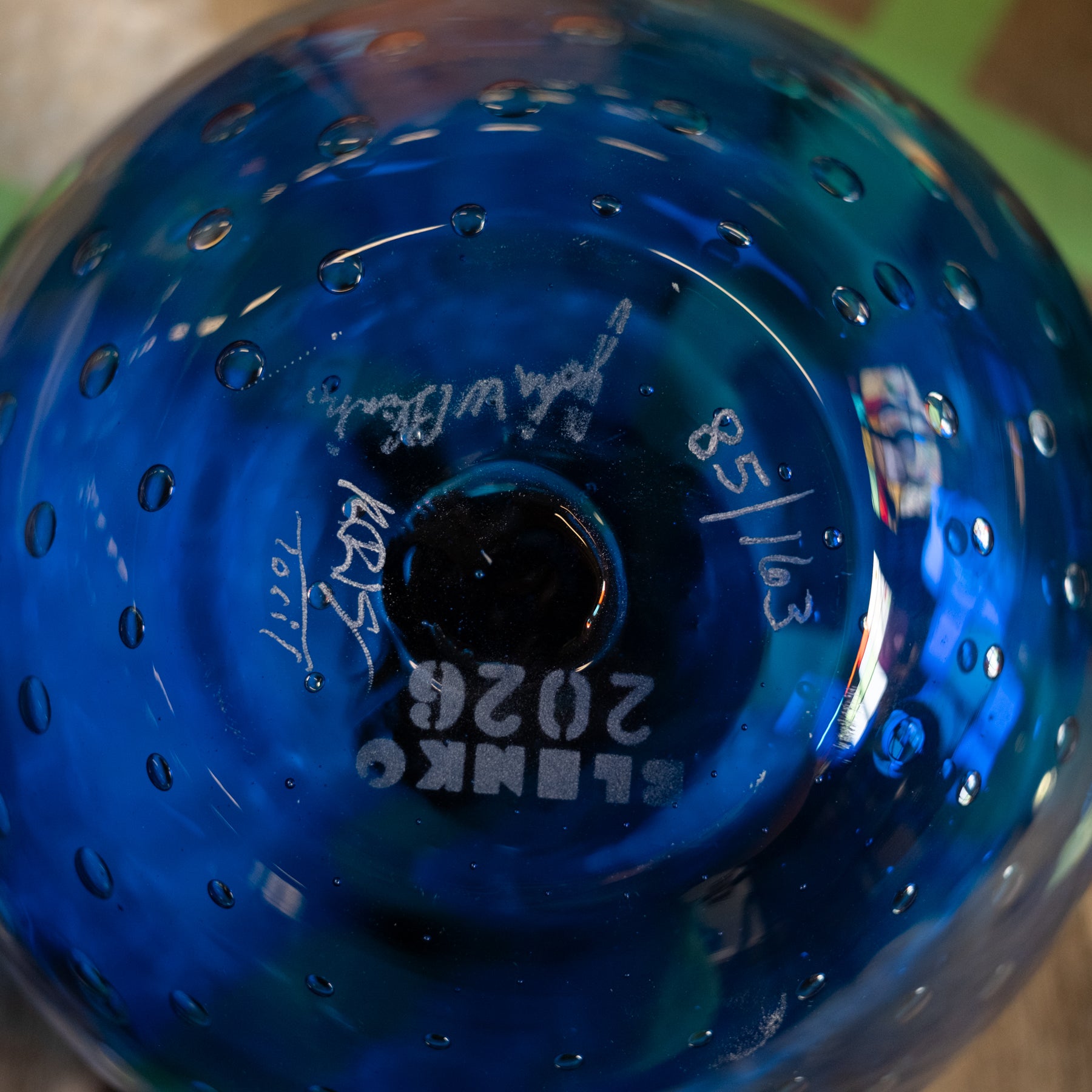 A close-up of a blue glass bowl or vase with a perforated bubble pattern, showing etched white text and numbers on the bottom including 'BLENK 2026' and '8/58'.