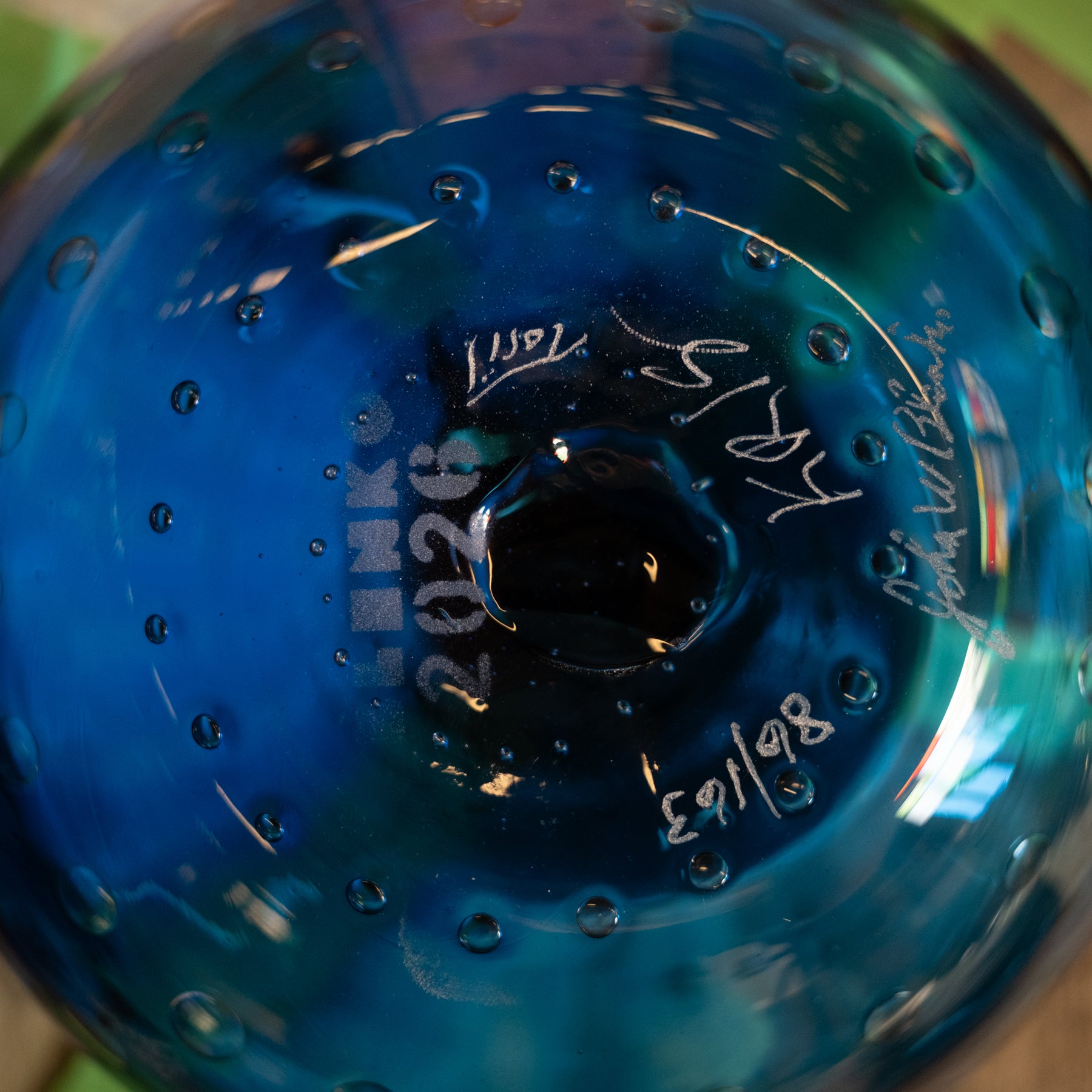 Close-up of the bottom of a blue glass bottle or vase with a central dark opening, showing embossed text, white handwritten markings, and small bubble-like glass details.