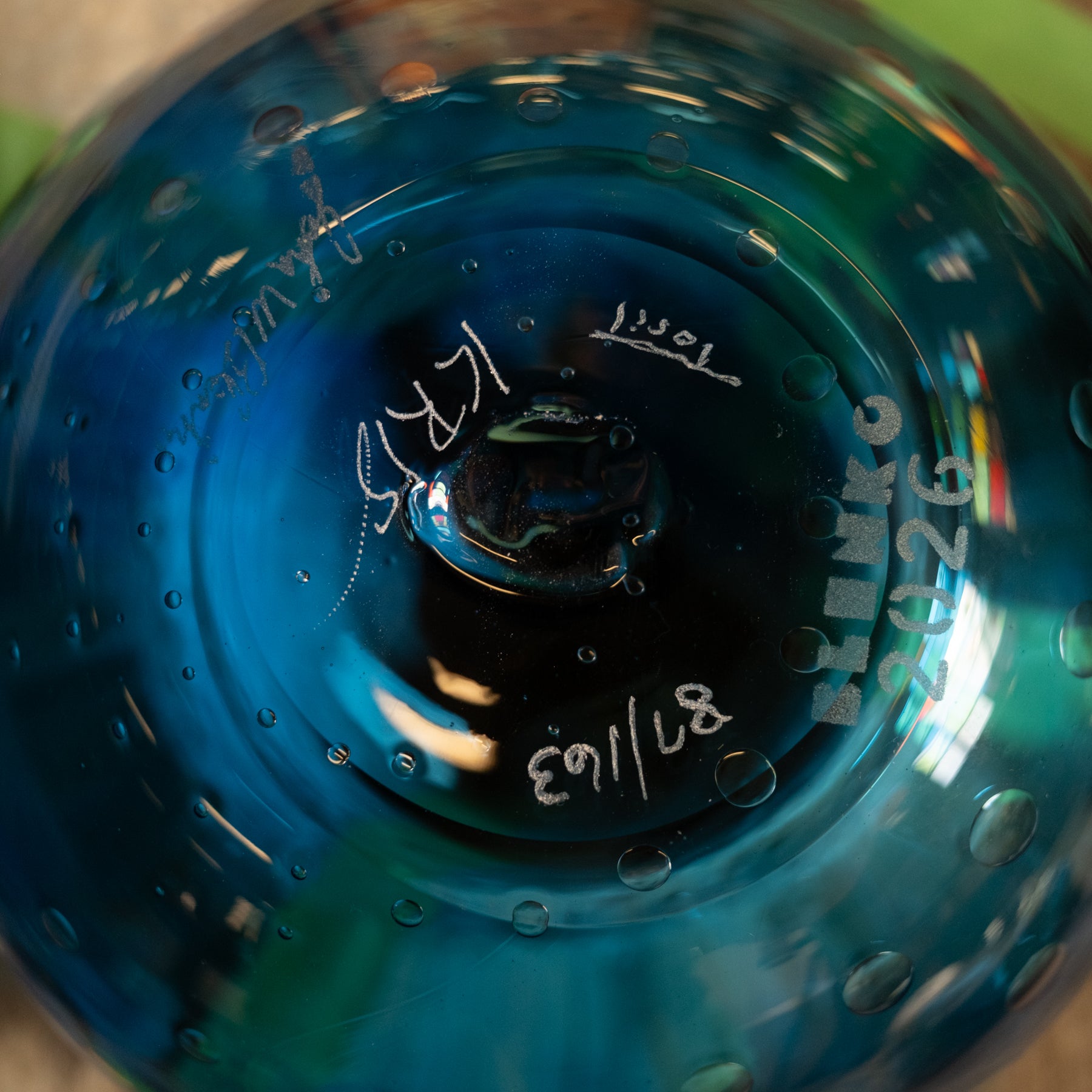 Close-up of the base of a blue glass bowl or vase with hand-written markings and bubble details, photographed against a blurred background.