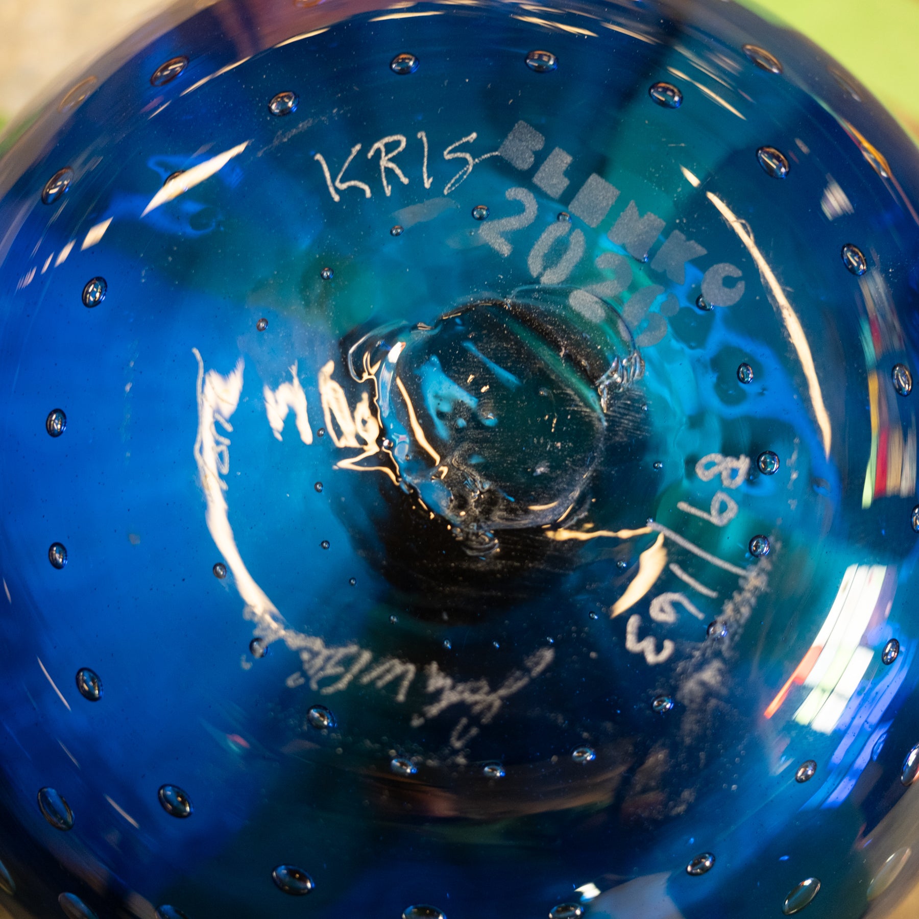 Close-up of the bottom of a transparent blue glass bowl or vase with bubble details, showing white handwritten markings and reflections.
