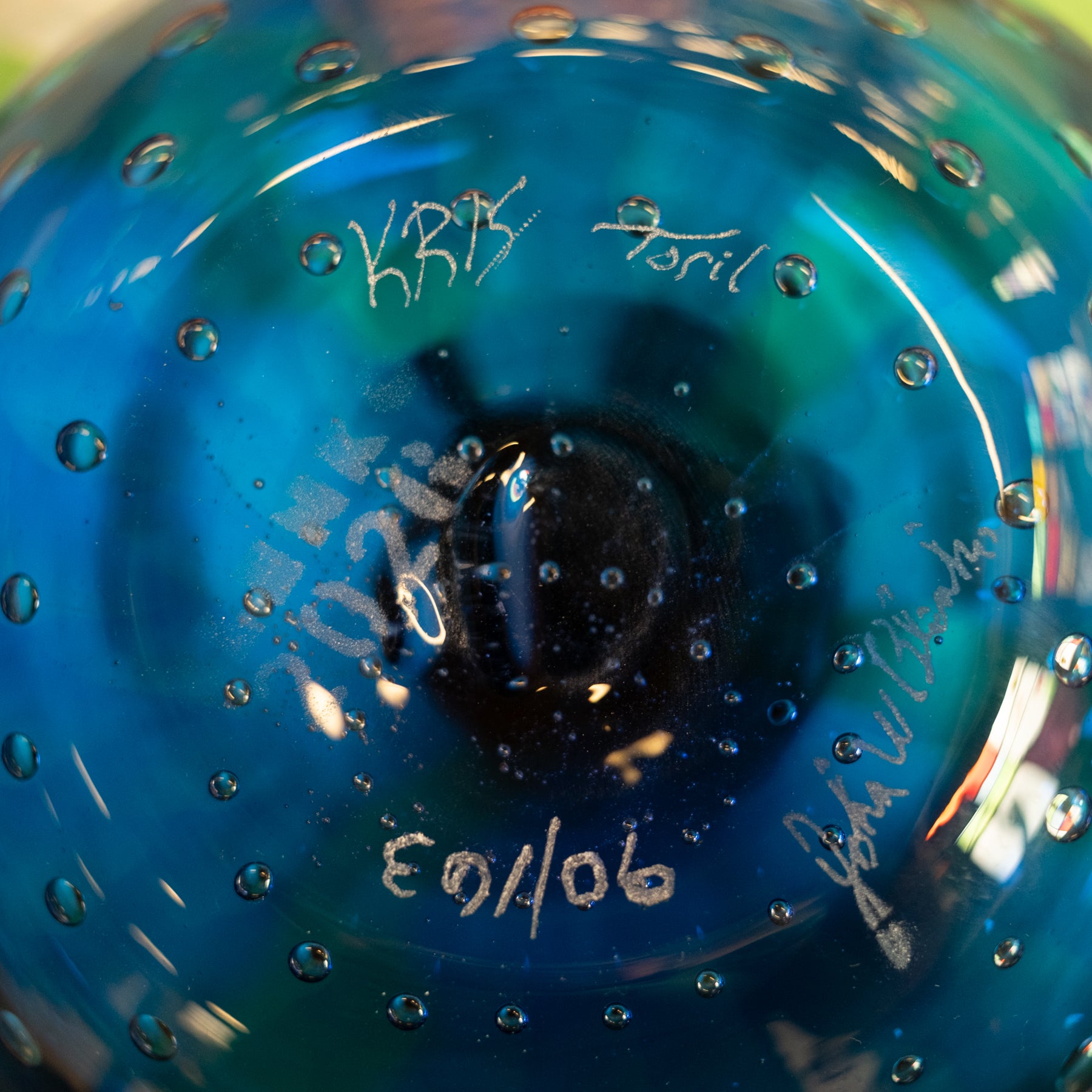 Close-up of the bottom of a blue glass bottle with embossed bubble details, featuring white handwritten signatures and a date '90/06/03'.